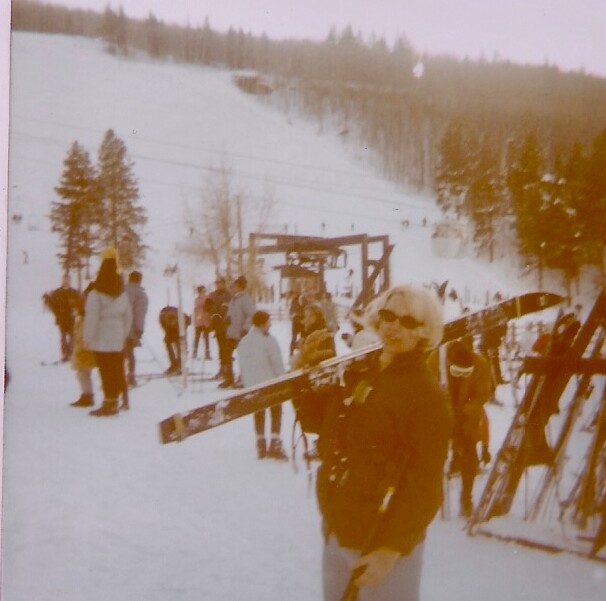 Person in sunglasses and a dark ski jacket carries a pair of skis over their shoulder on a snowy ski resort, with other skiers and a lift in the background.