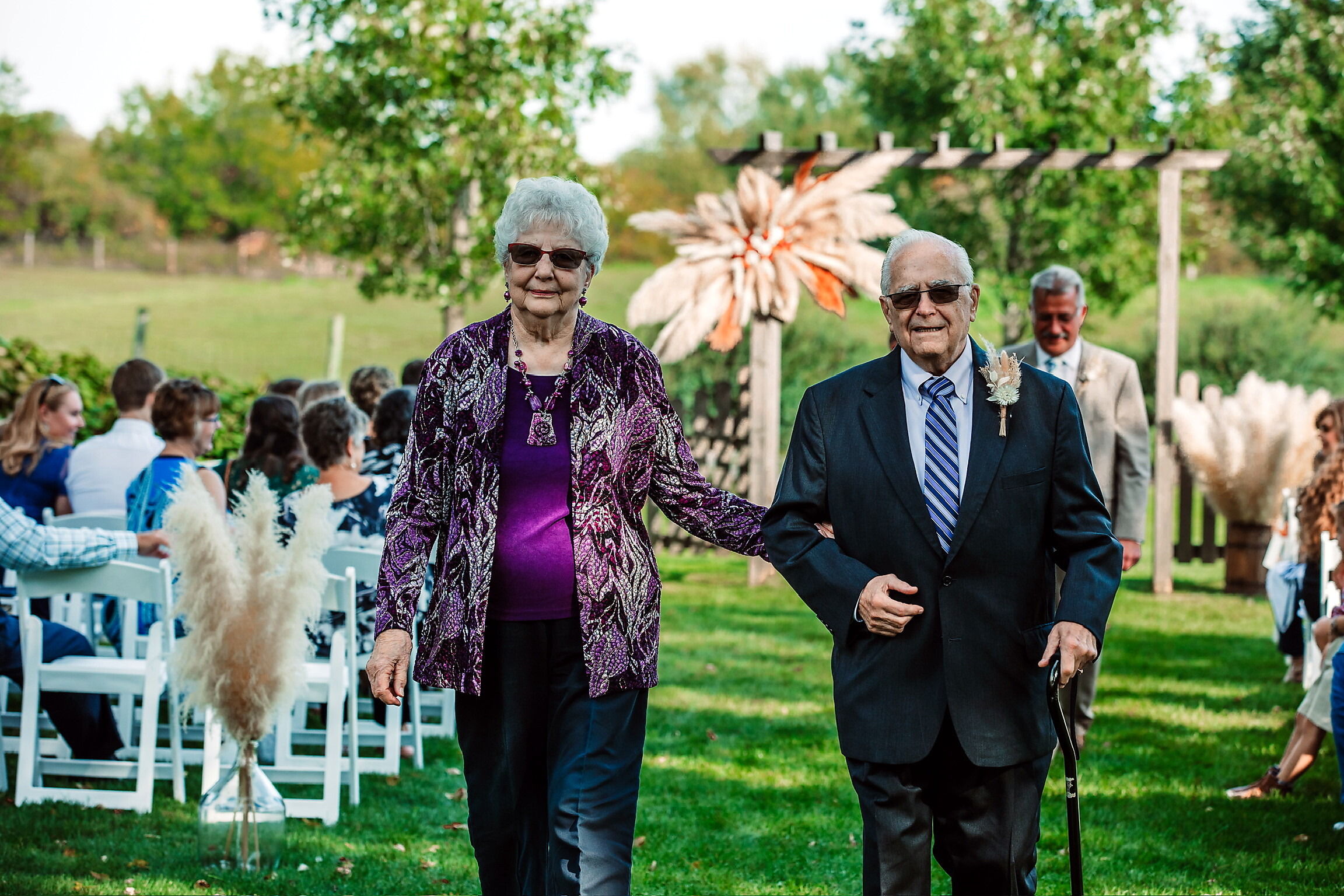Older couple walking arm-in-arm down an outdoor ceremony aisle, guests seated on white chairs to the side under green trees.