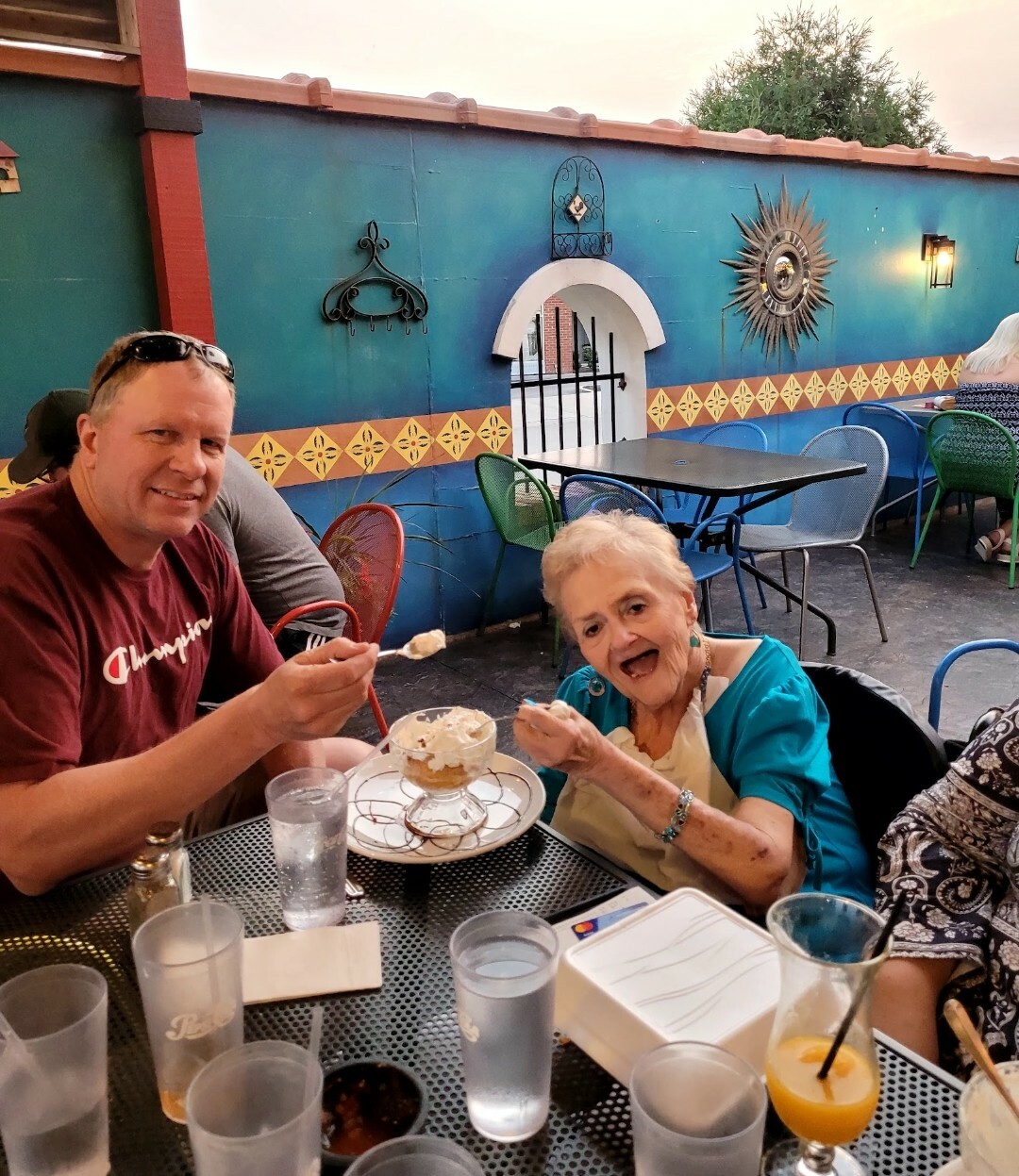 Man and an elderly woman enjoy ice cream sundaes at an outdoor cafe, smiling at the camera.