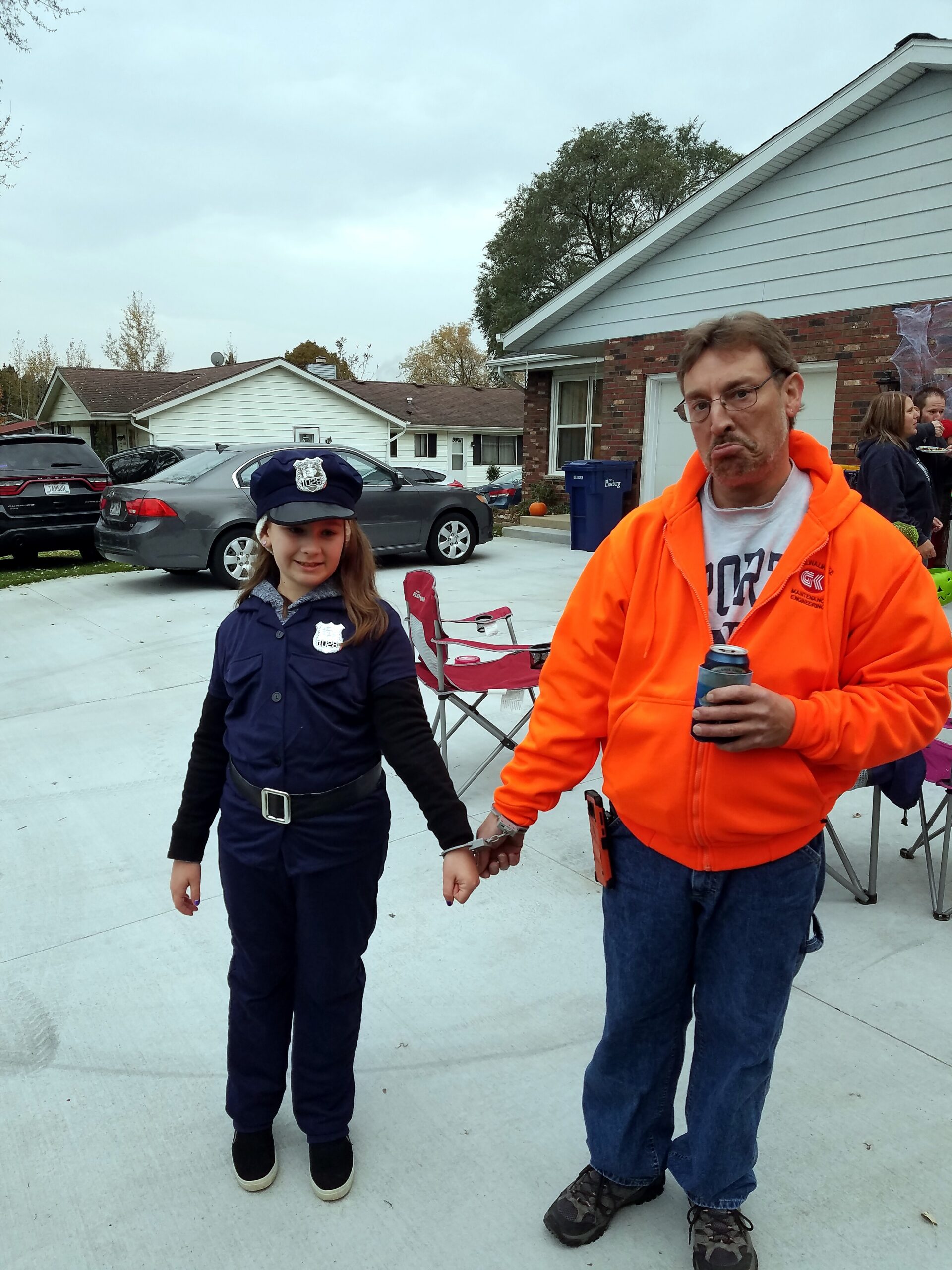 Man in a bright orange hoodie holding a beverage, standing beside a young girl in a police costume in a suburban driveway scene with houses in the background on a cloudy day.