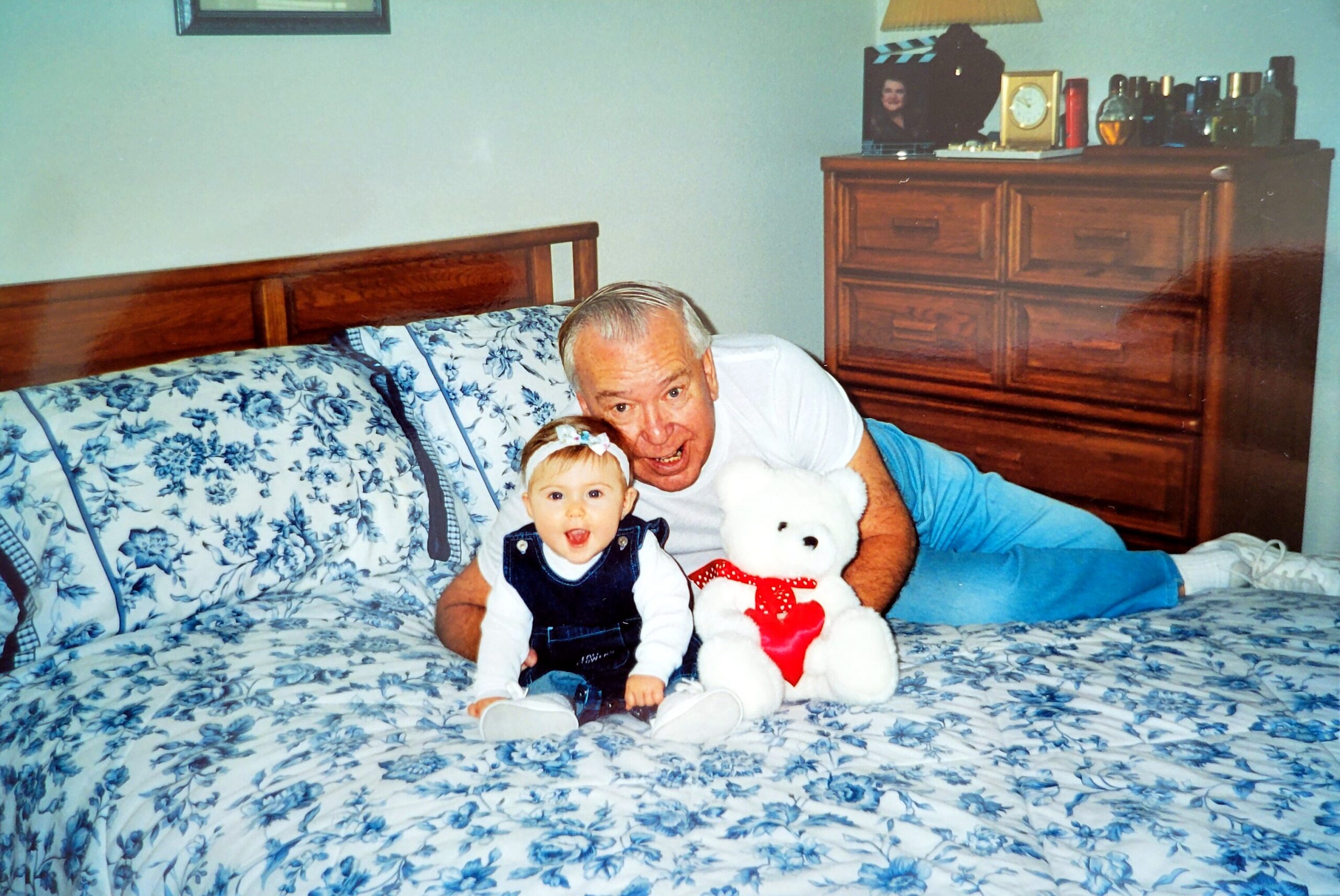 Older man lying on a bed with a baby and a white teddy bear; blue floral quilt and a wooden dresser in the background.