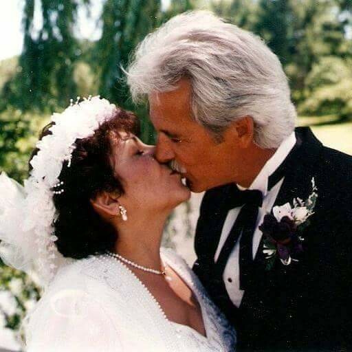 Bride and groom kiss outdoors on their wedding day; bride wears a veil and pearl necklace, groom in a tuxedo with a boutonniere.