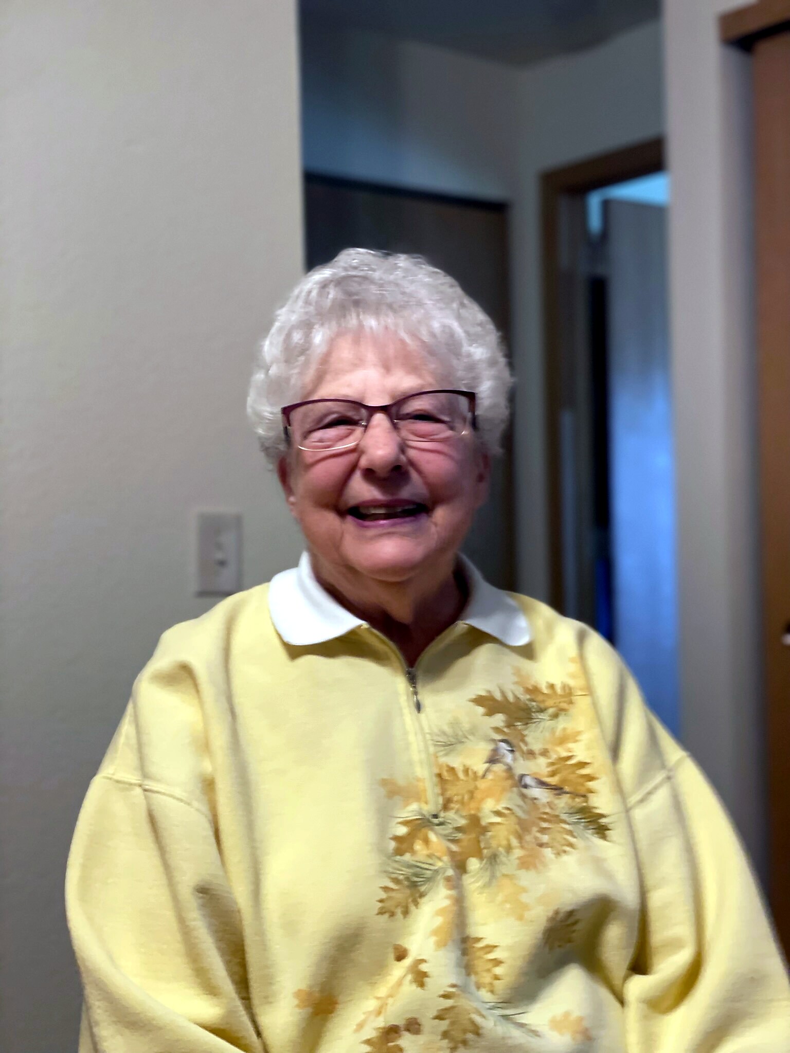 Smiling elderly woman with short white hair and glasses, wearing a yellow floral-patterned sweater indoors.