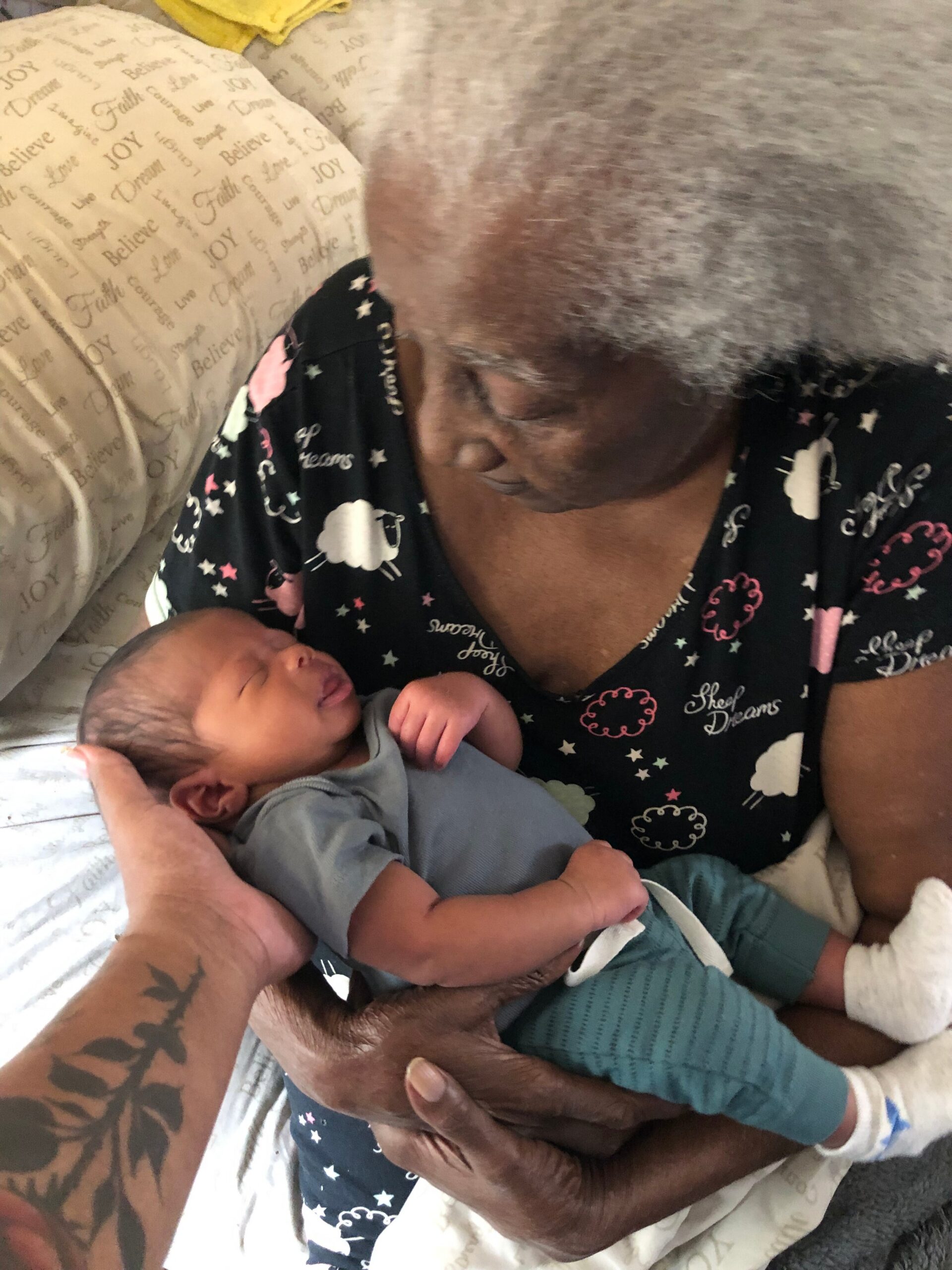 Adult with a tattooed forearm gently cradling a sleeping newborn baby in a blue onesie.