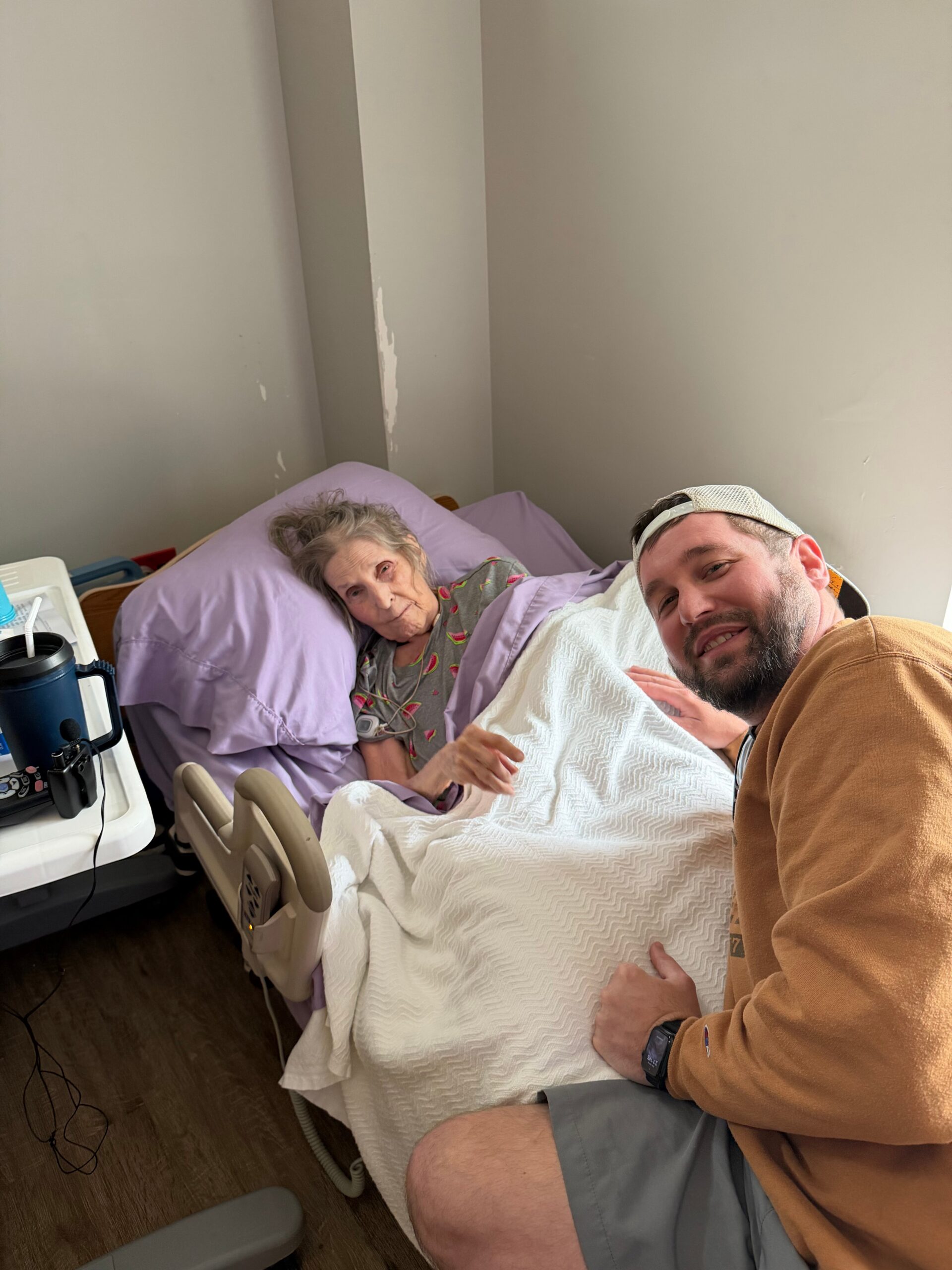 Man taking a selfie beside an elderly woman in a hospital bed, she rests with a purple pillow and white blanket, bedside medical equipment nearby.