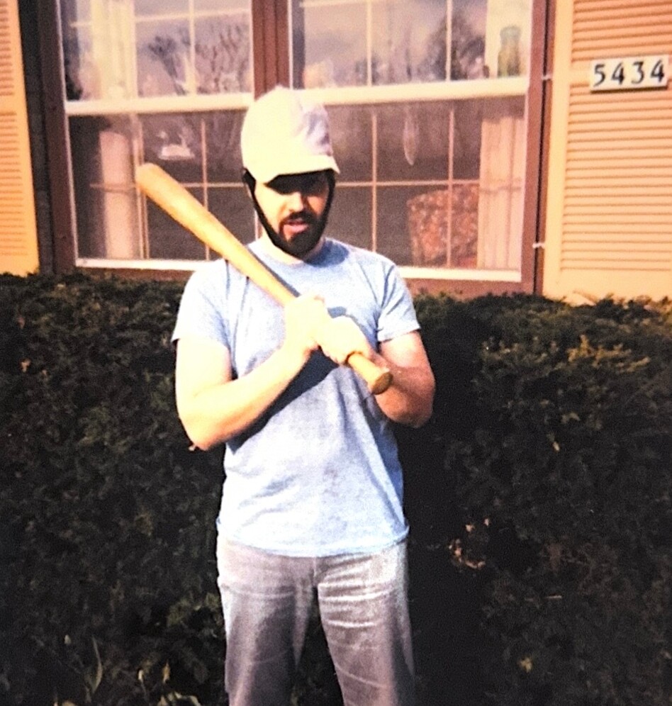 Bearded man wearing a white helmet and light blue shirt, holding a wooden baseball bat across his chest outdoors.