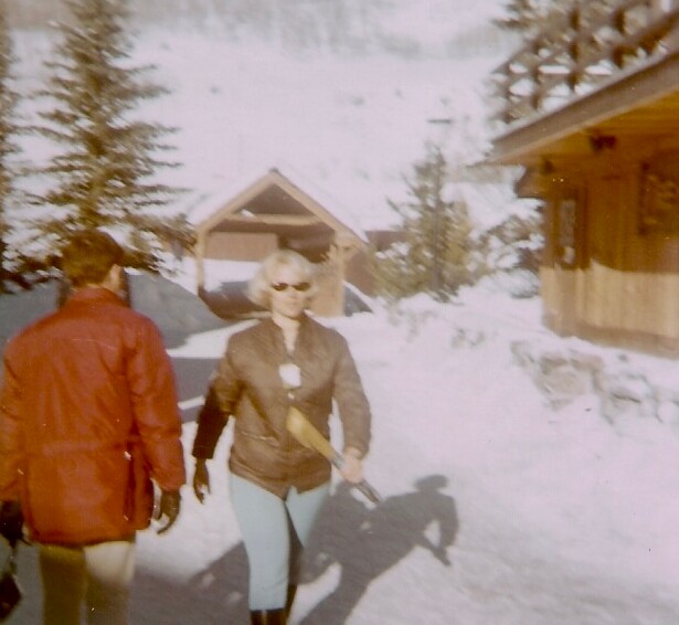 Two people walk through a snowy resort area with cabins in the background; a woman in a brown jacket and light jeans leads, while a person in a red jacket follows.