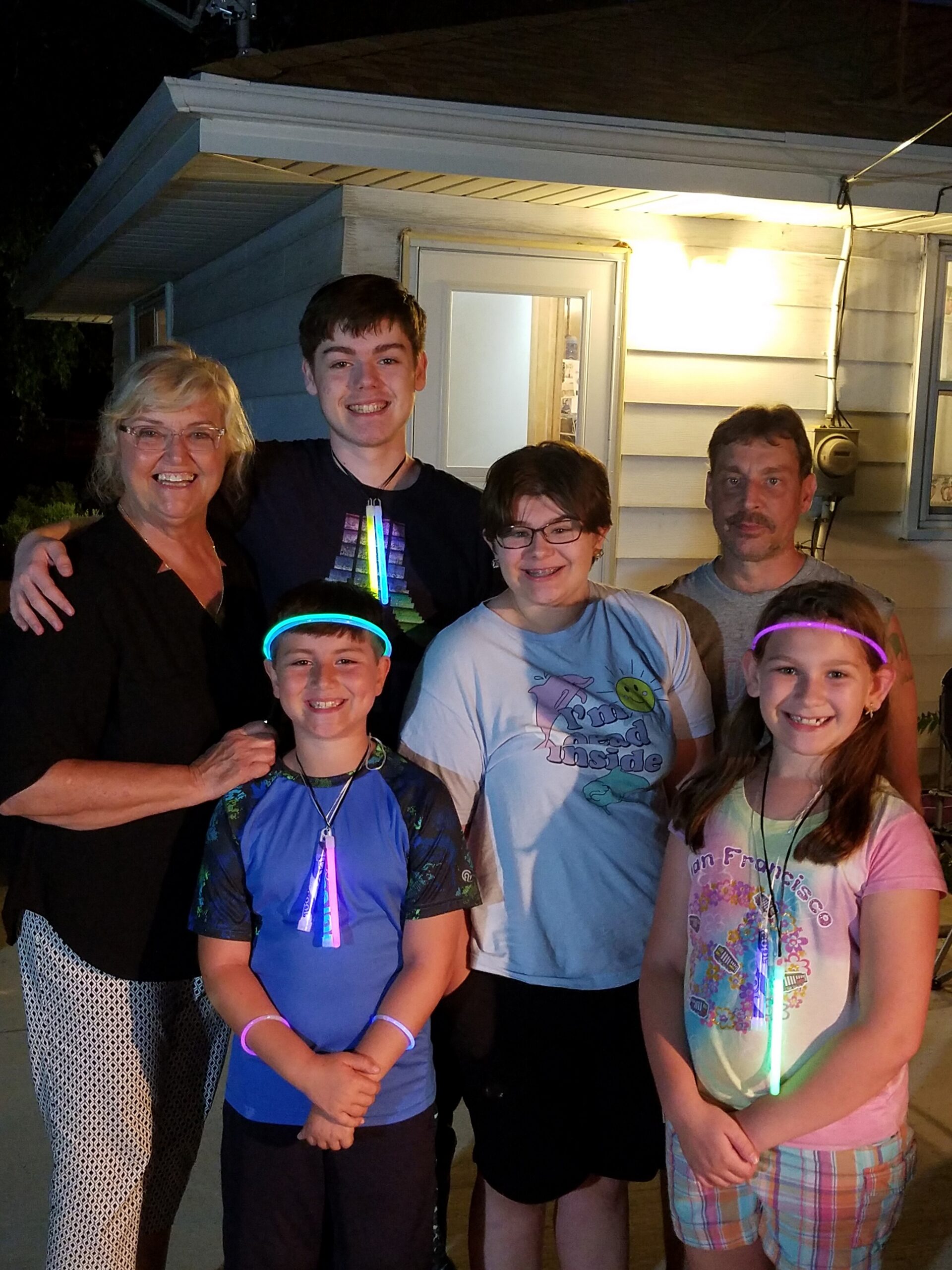Smiling multi-generation family posing together outside a house at night, with glow necklaces and bracelets glowing in the dark.