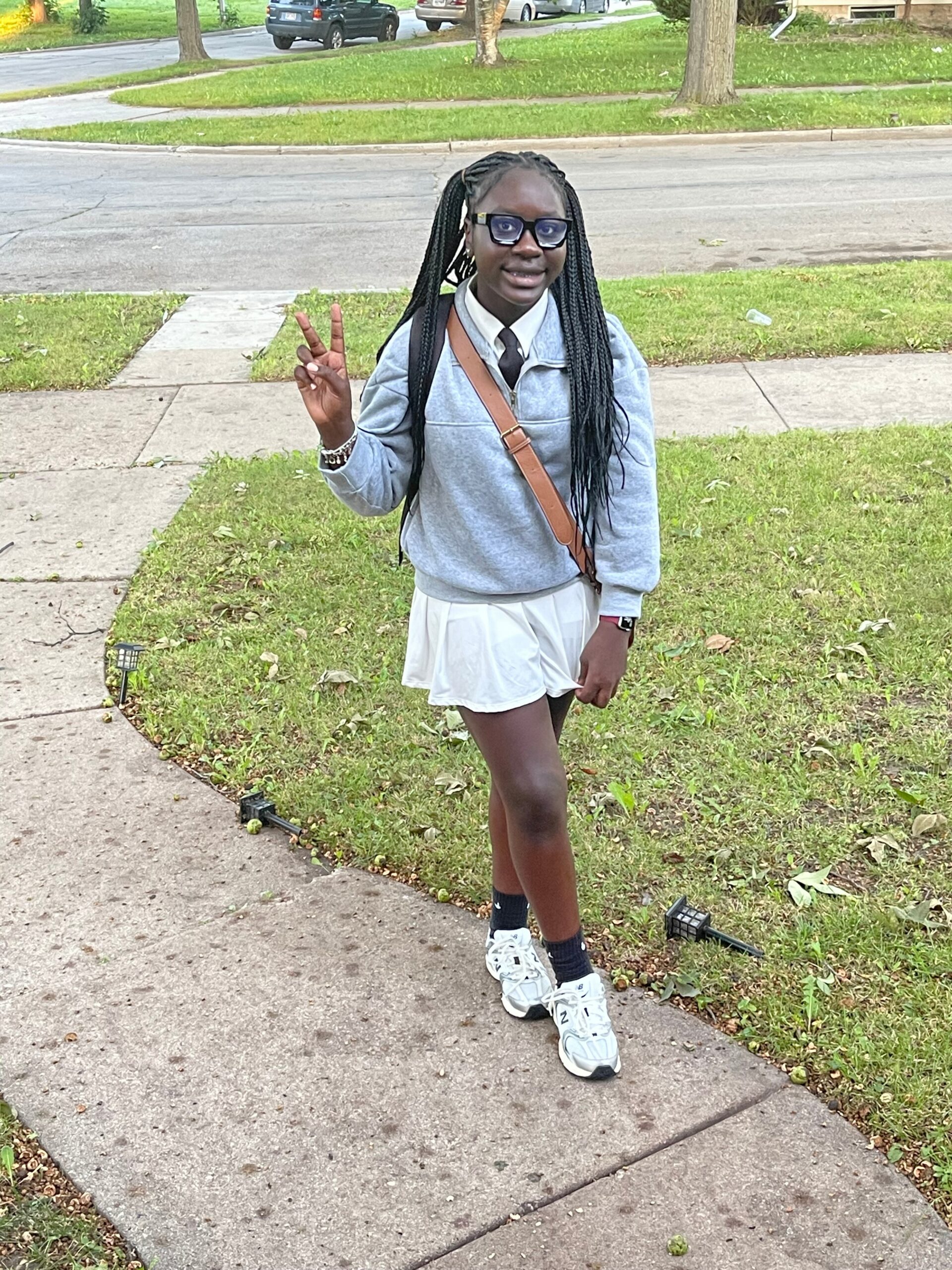 Girl standing on a sidewalk outdoors, making a peace sign with her right hand, wearing a gray sweatshirt, white skirt, black tie, glasses, and a brown crossbody bag.