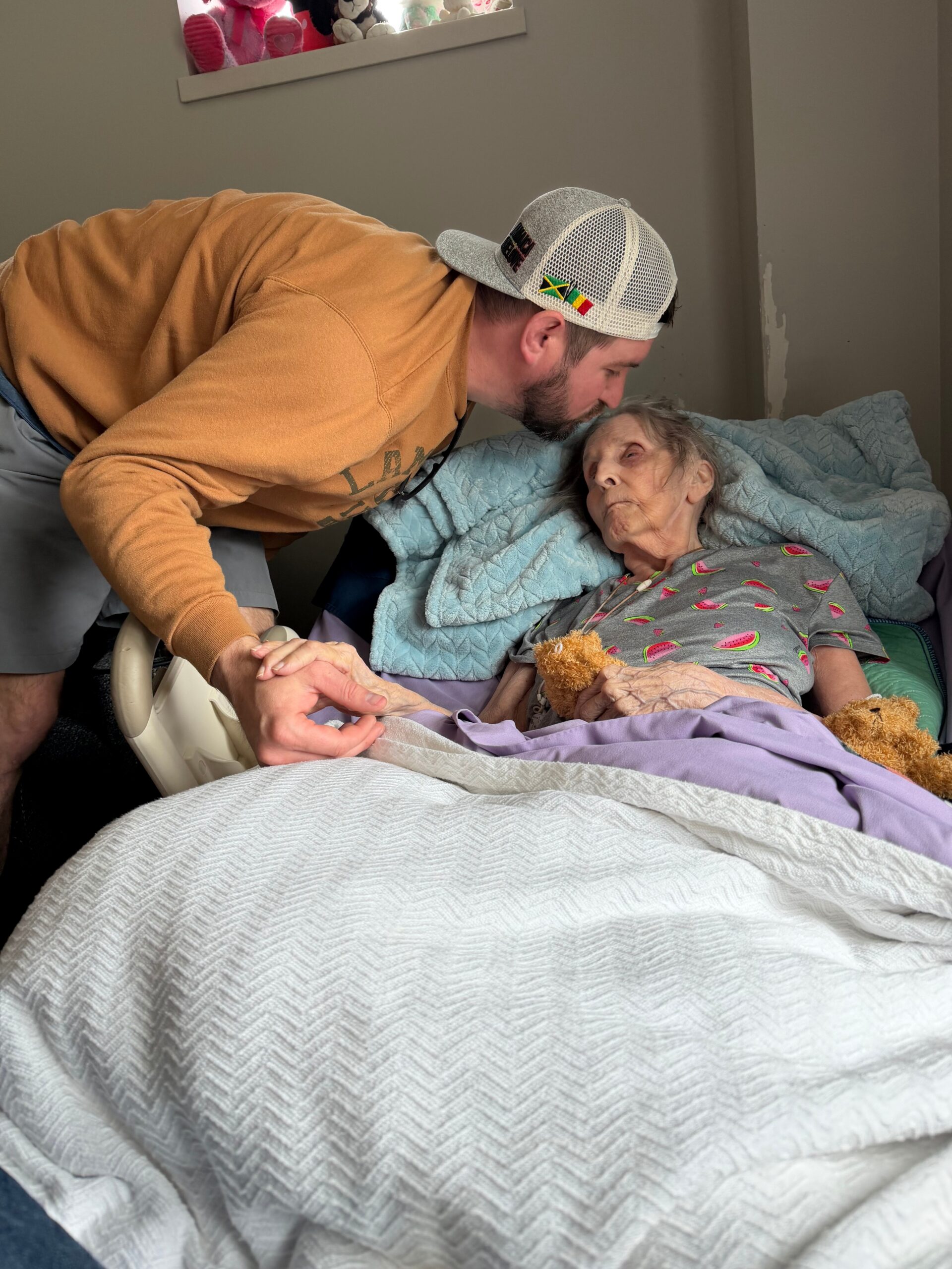A caregiver leans over an elderly woman in bed, holding her hand in a comforting gesture.