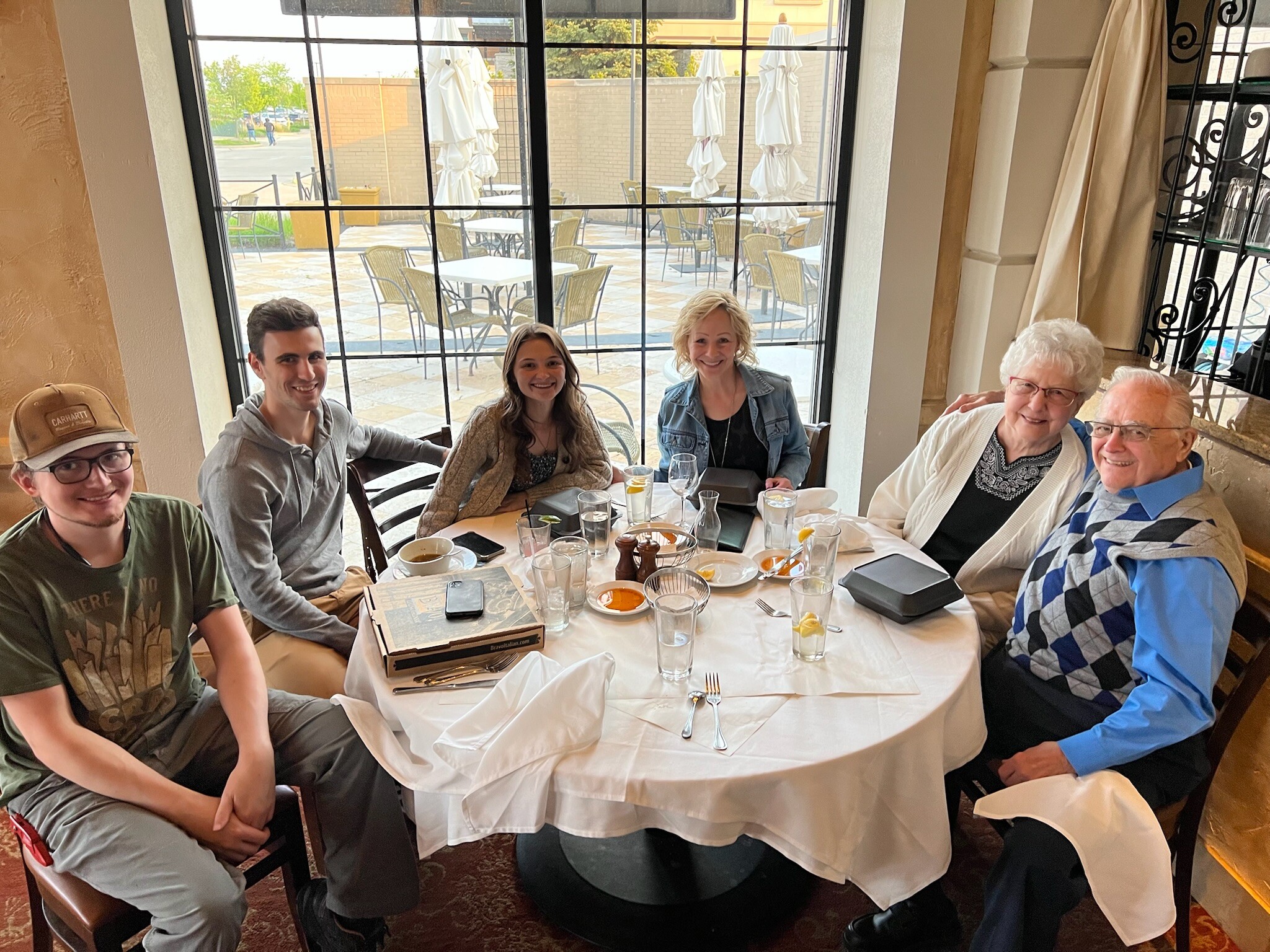Six people smiling around a round table in a restaurant, with drinks and plates on the table and a large window behind them.