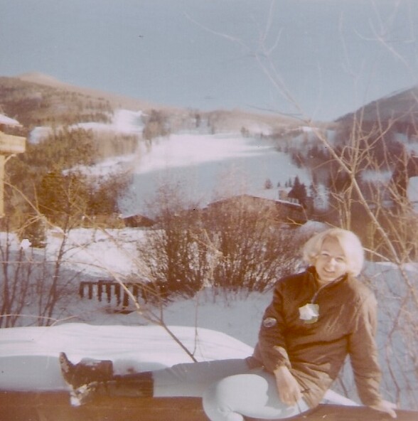Smiling woman in a brown jacket sits on a snow-covered surface with a snowy valley, hills, and a lake in the background.