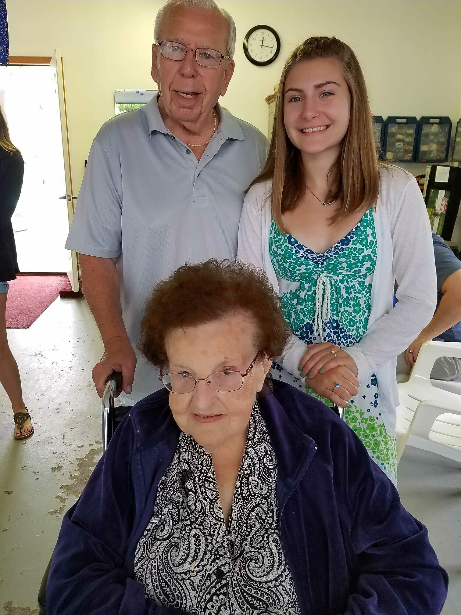 An elderly woman in a wheelchair in the foreground, with a smiling young woman standing behind her in a bright room.