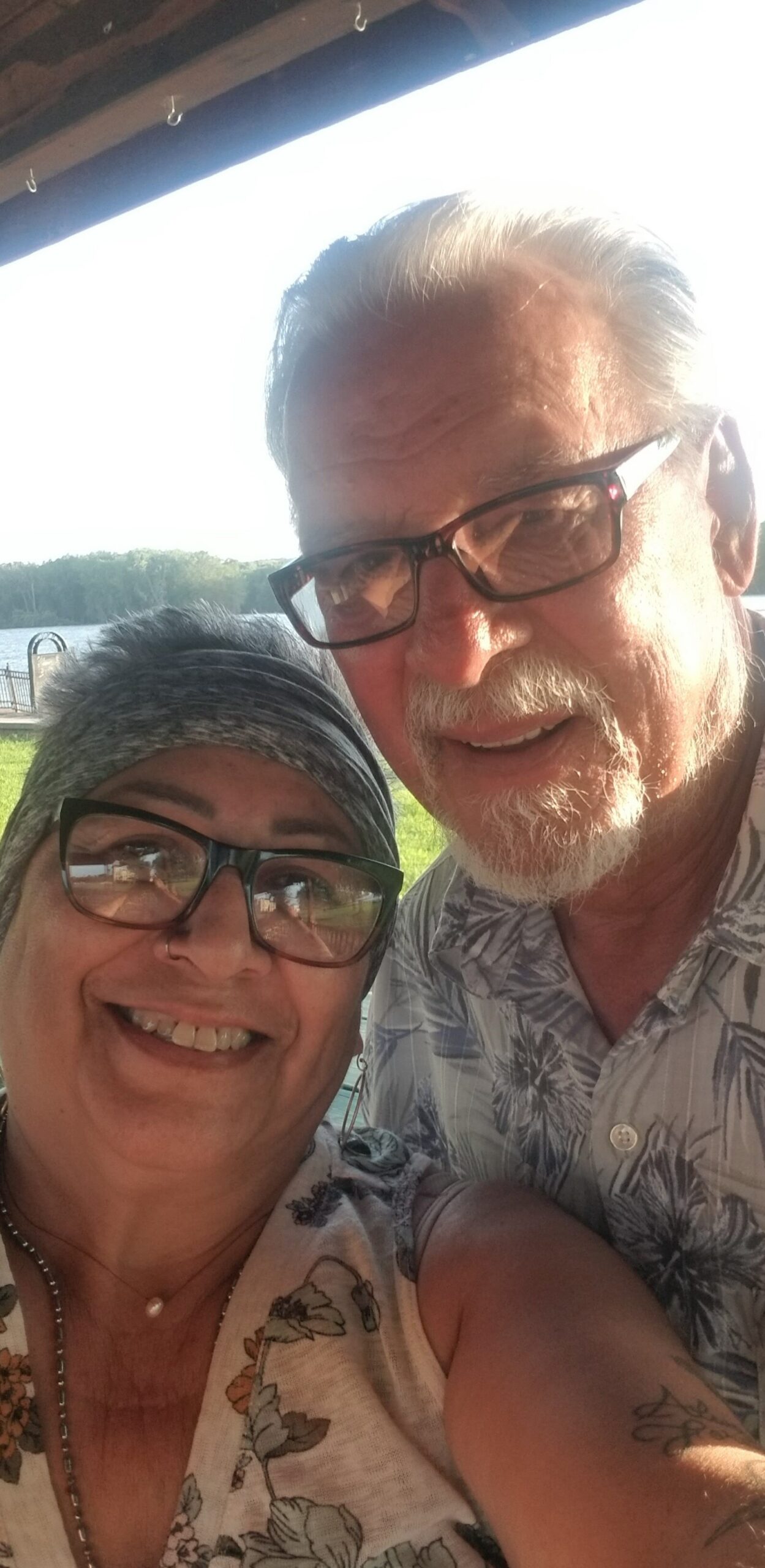 Two people smiling for a selfie outdoors, a man with gray hair and glasses beside a woman wearing a headscarf and glasses by a lake park.