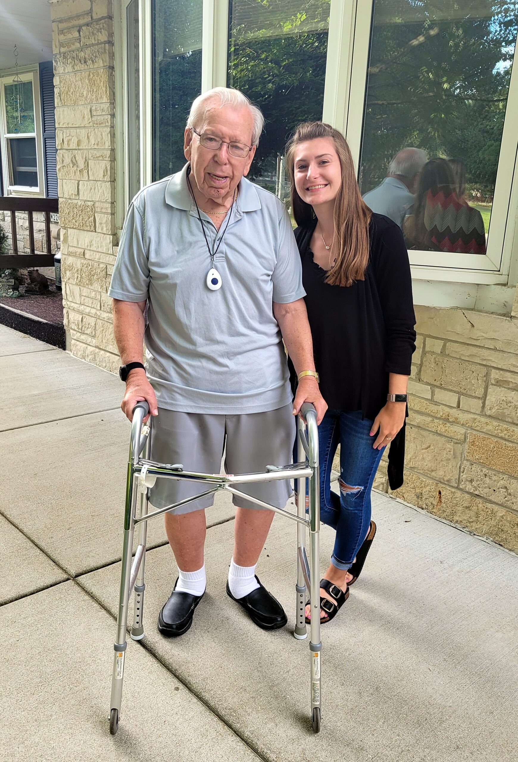 Elderly man with a wheeled walker on a sidewalk, smiling, with a younger woman standing beside him outside a stone house.
