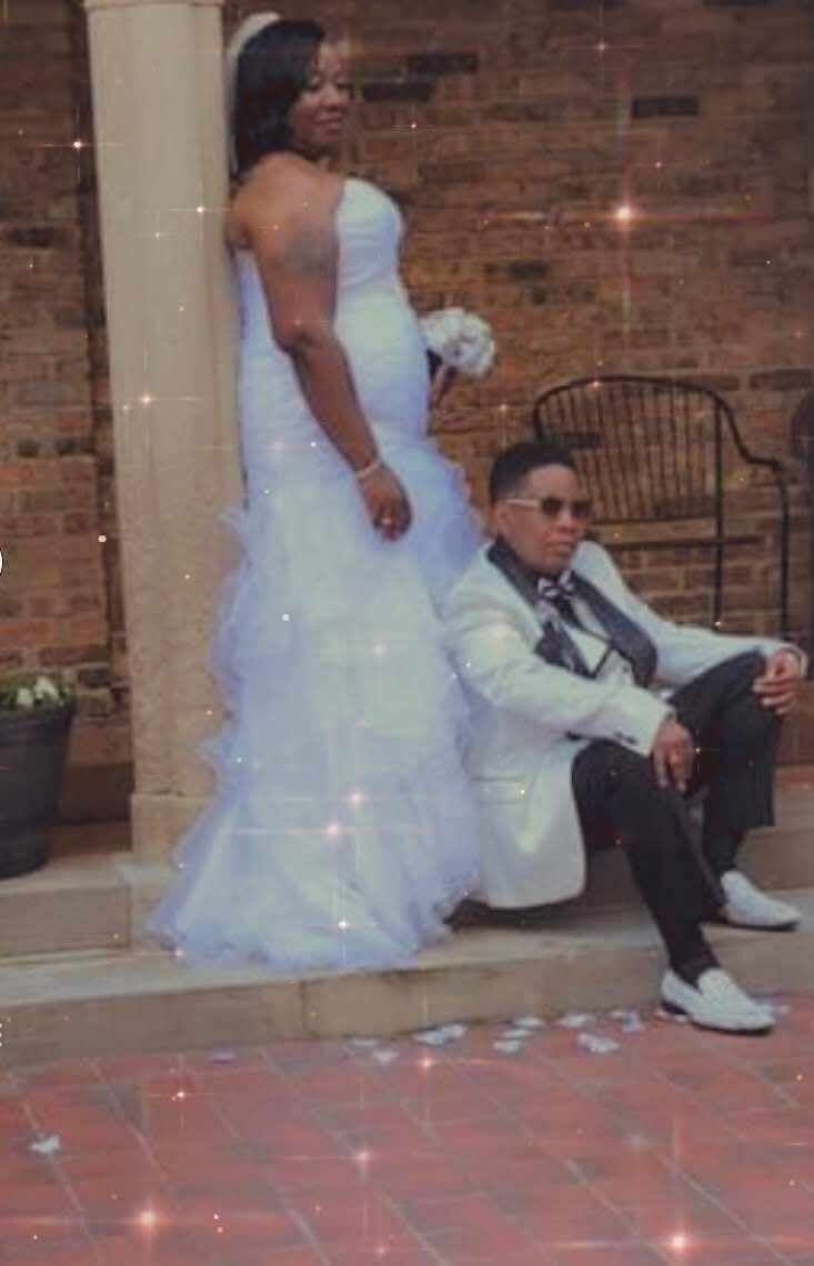 Bride in a strapless white wedding gown stands beside a man in a light suit seated on steps, holding a bouquet.