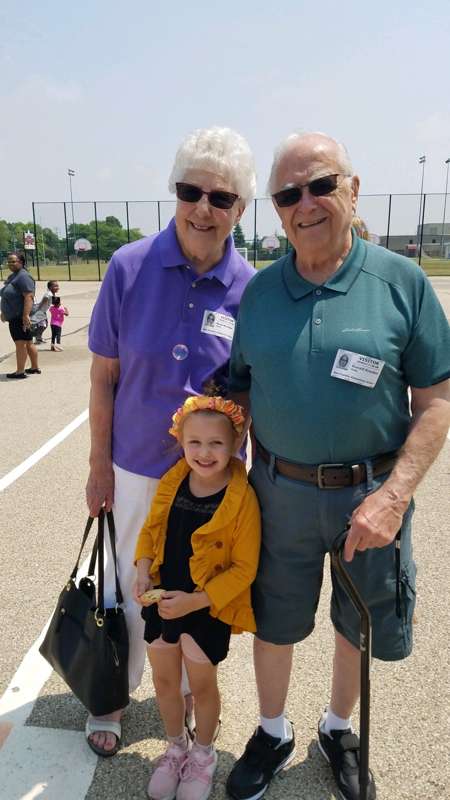 Smiling elderly couple with a young girl at an outdoor event, standing by a basketball court on a sunny day; badges visible on their shirts.