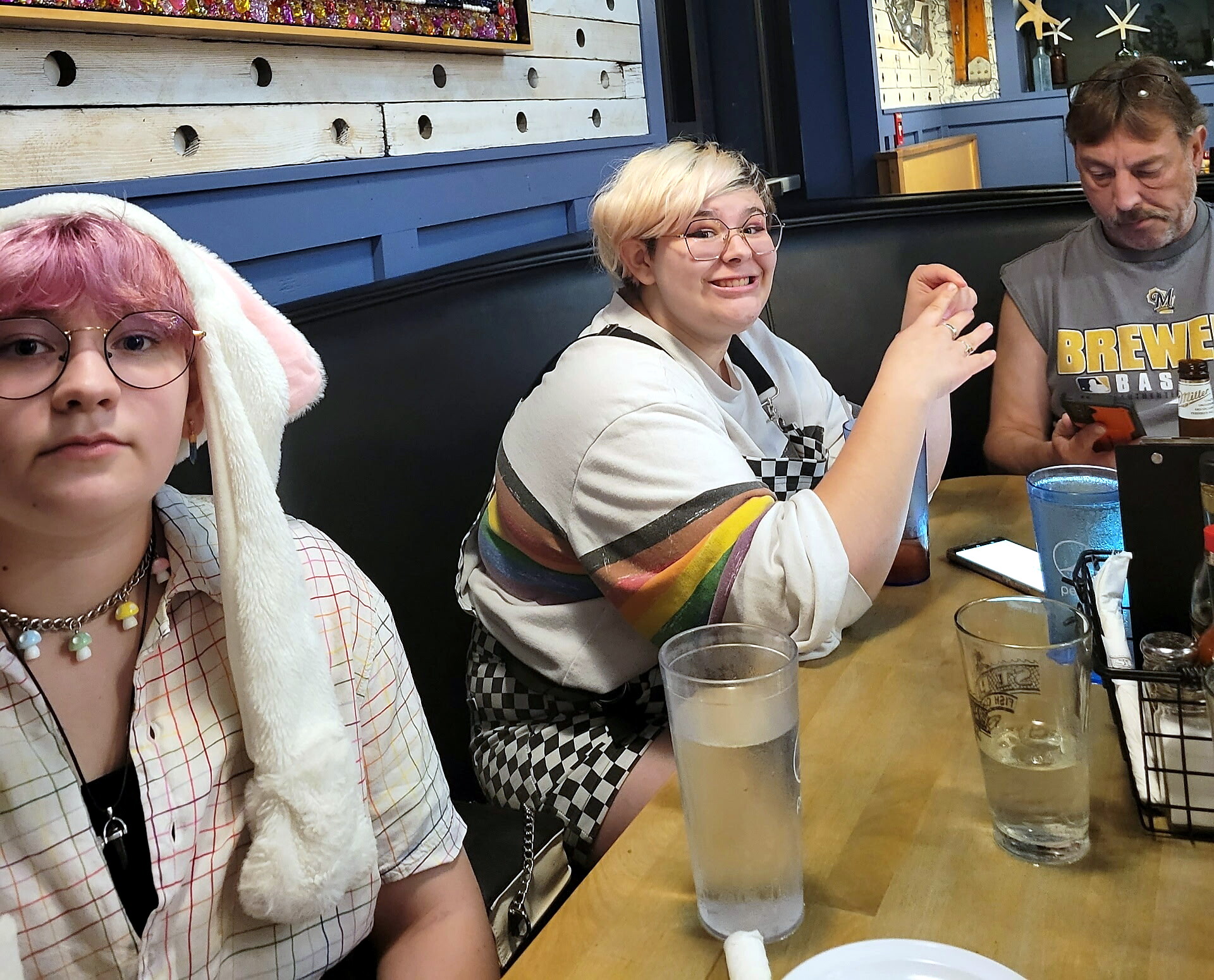 Three friends at a restaurant table with drinks; left person wears bunny ears, center person smiles at camera, right person looks at phone.