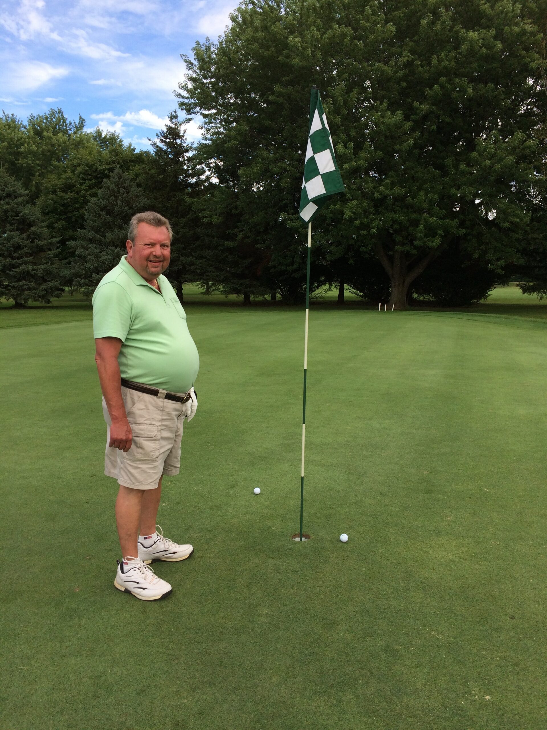 Smiling man in a light green polo and khaki shorts stands on a golf green beside a striped flagstick and hole, with two golf balls nearby and trees in the background.