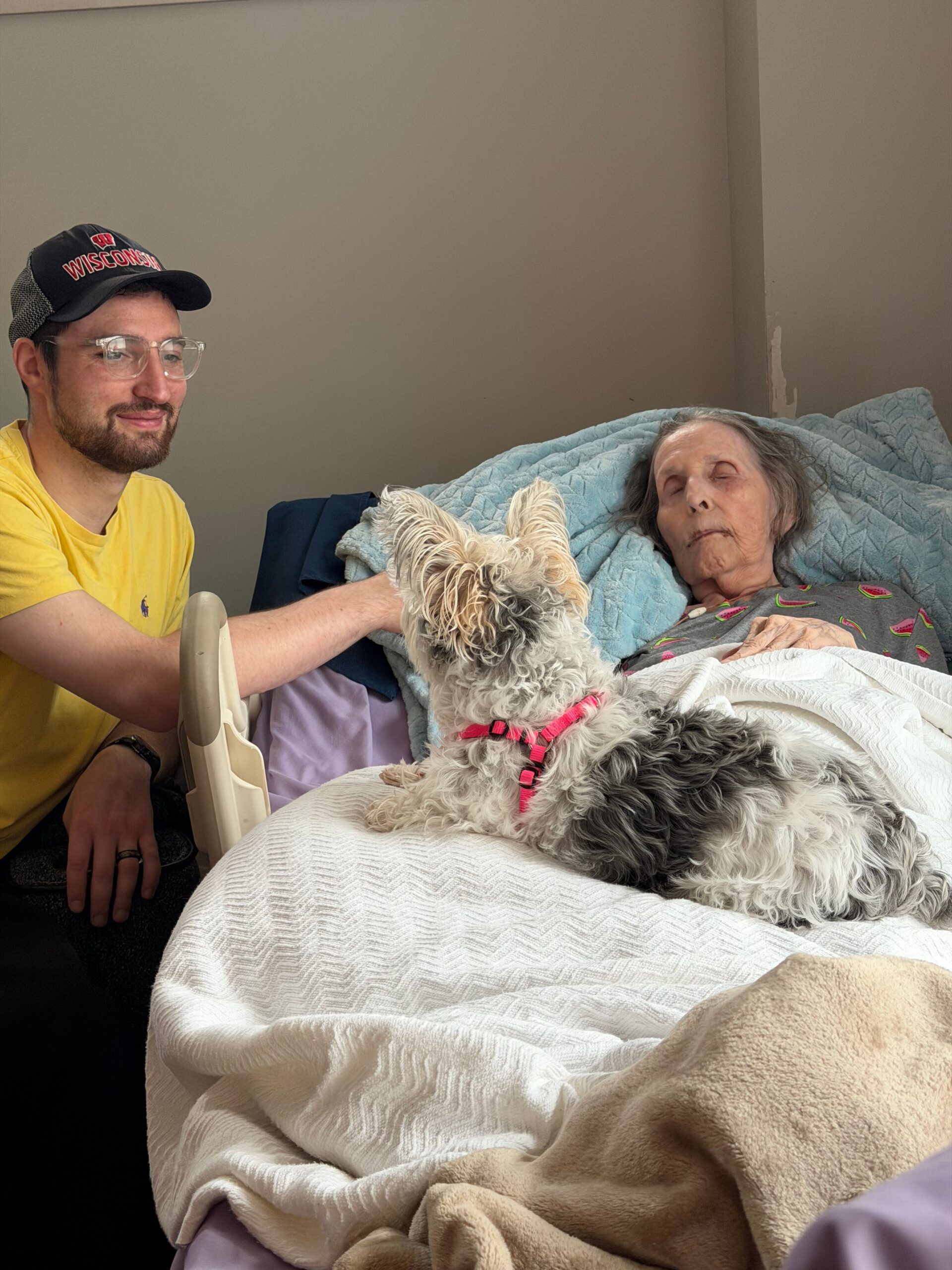 Caregiver wearing a Wisconsin cap sits beside a bed, petting a fluffy dog next to an elderly woman resting in blankets.