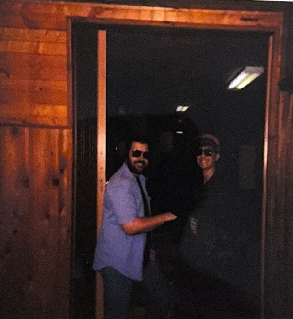 Two men wearing sunglasses stand in a wood-paneled doorway at night, one in a light blue shirt and the other in dark clothes, smiling at the camera.