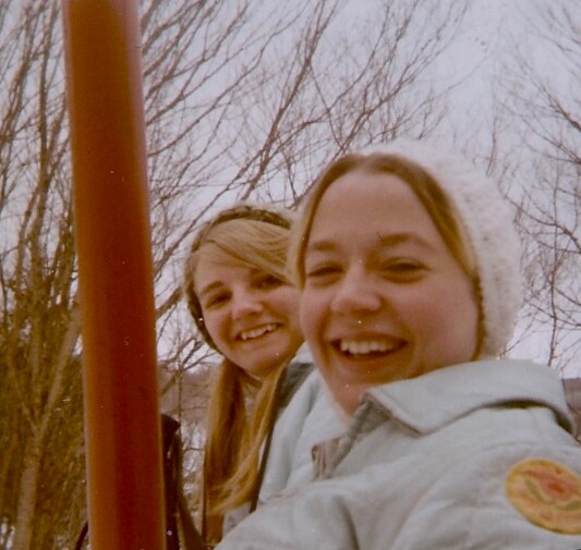 Two smiling women posing outdoors in winter; one wearing a white knit hat and light jacket, near a red pole.