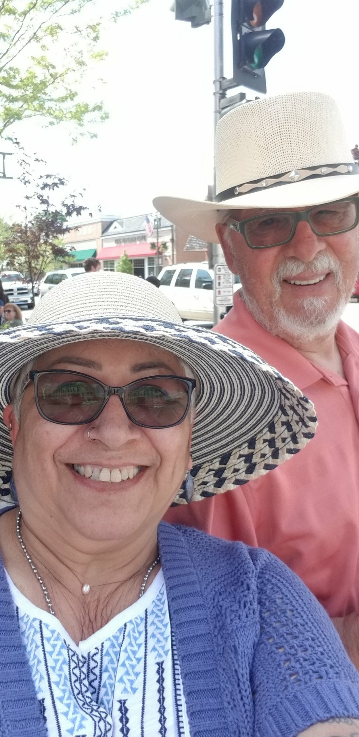 Smiling woman in a striped sunhat and glasses taking a selfie with a man behind her in a cream straw hat and pink shirt on a sunny street.