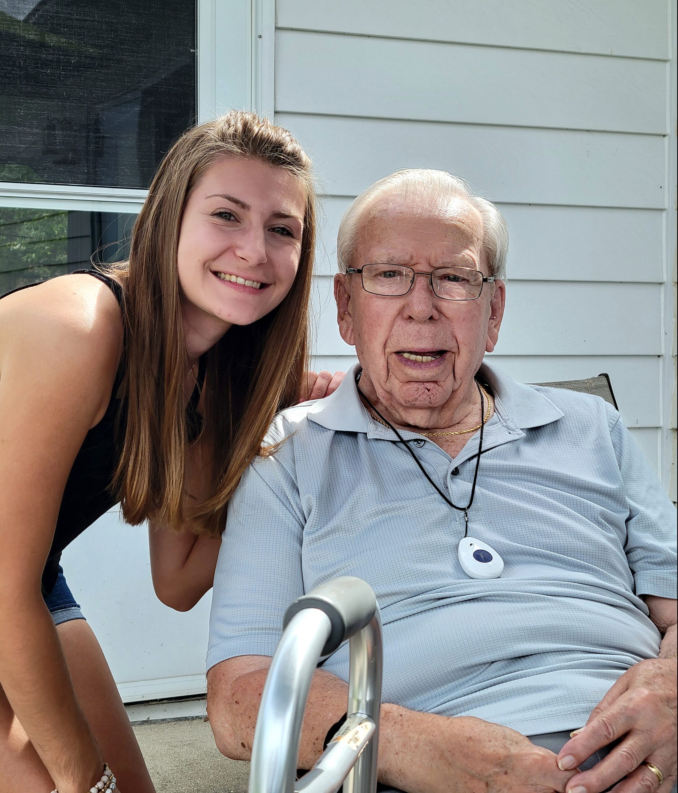 Young woman with long hair smiling beside an elderly man in a light polo shirt sitting in a wheelchair on a porch area outside a house with light siding.