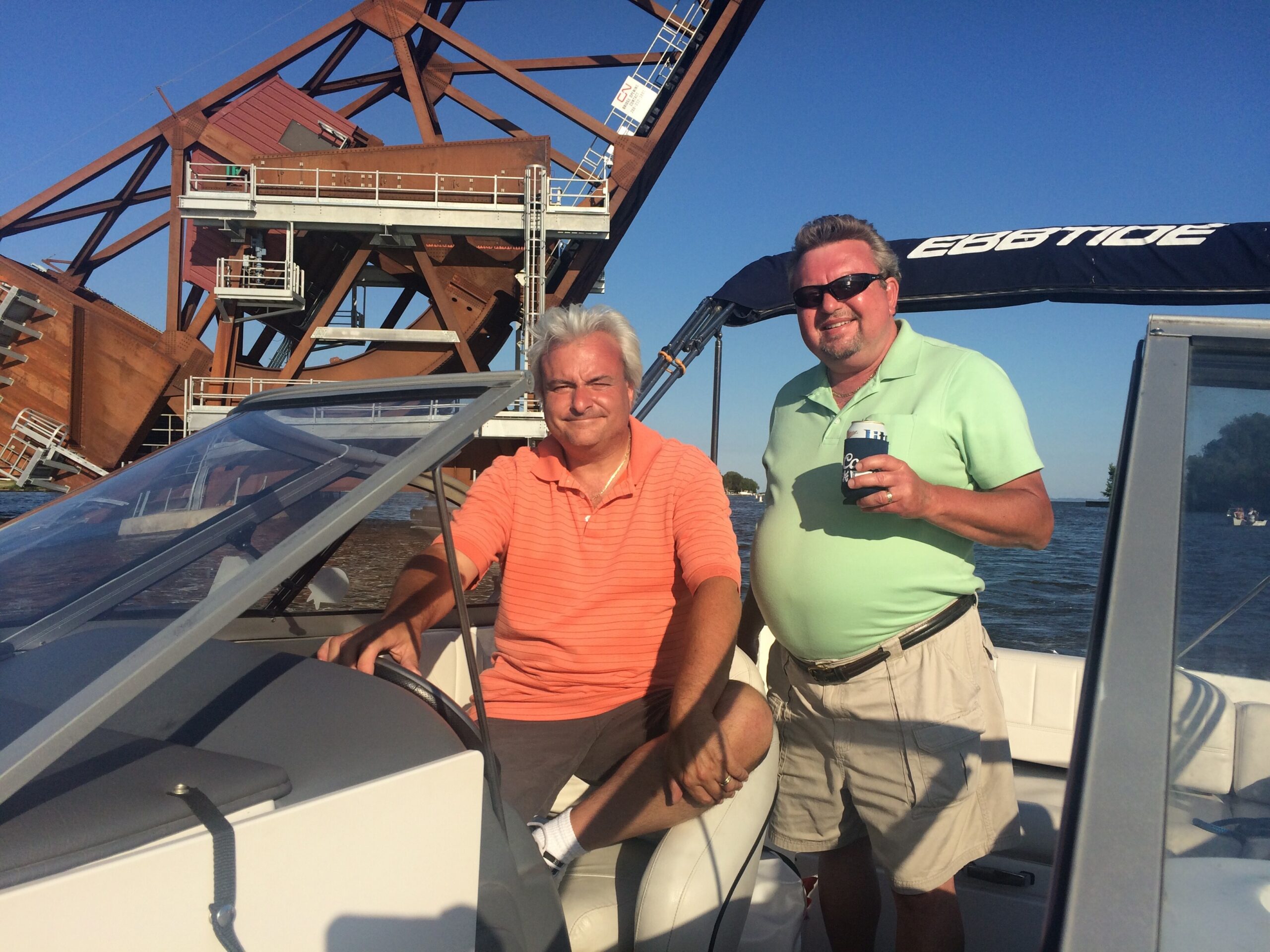 Two men stand at the helm of a boat, one in an orange polo and the other in a light green shirt with sunglasses, holding a can of drink against a backdrop of a large rusted crane and blue sky.