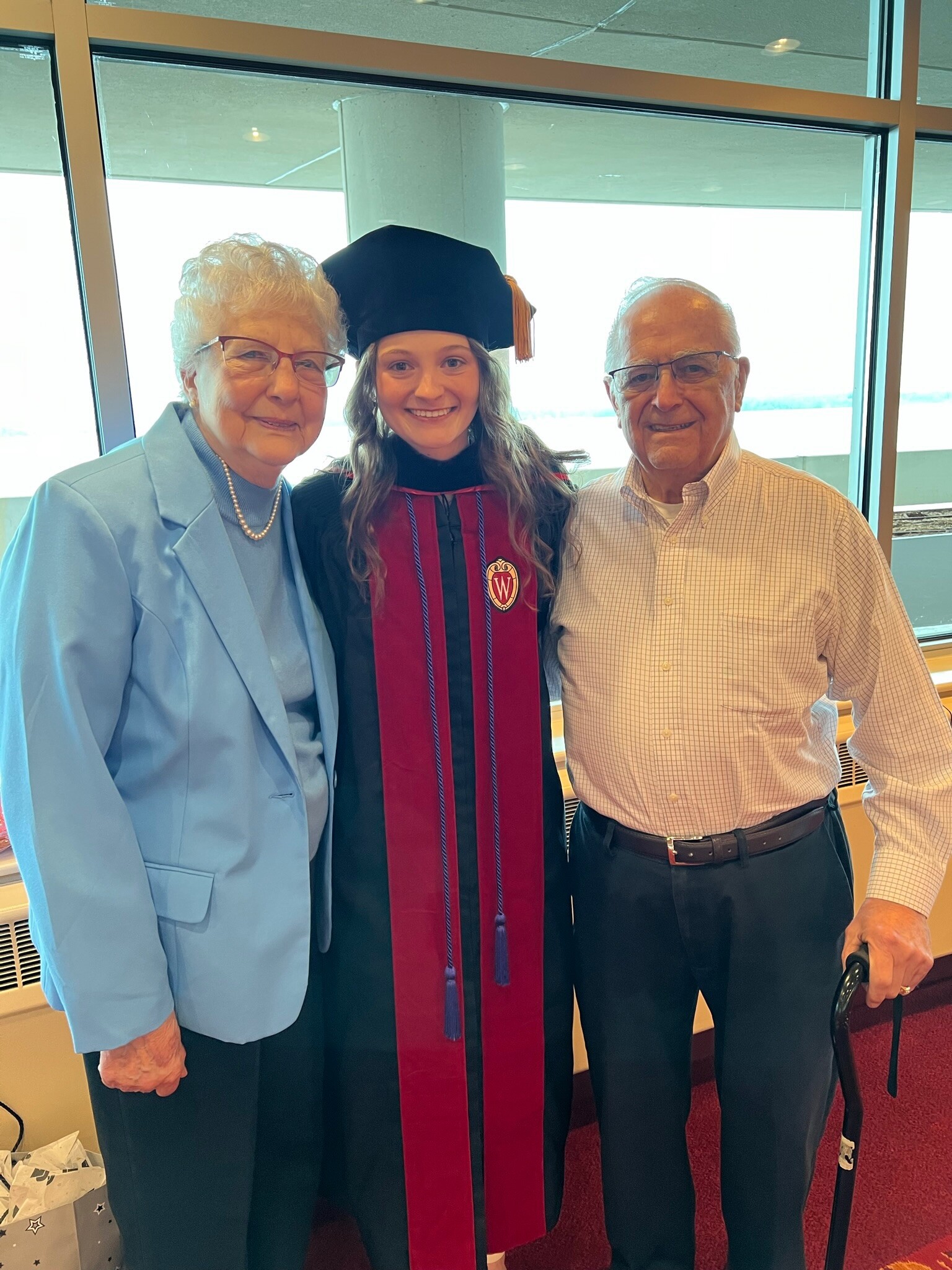 Three people pose indoors: a smiling grandmother in a light blue blazer, a graduate in a black cap and maroon gown, and a smiling grandfather in a plaid shirt.