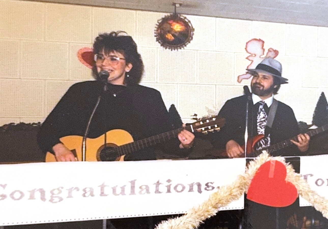 Two musicians performing on a stage: a woman with an acoustic guitar sings into a microphone, a man in a hat plays guitar beside her.