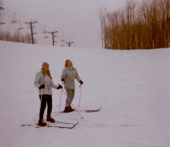 Two skiers in light jackets standing on a snowy slope with ski poles and skis on, chairlift in the background and bare trees lining the hill.