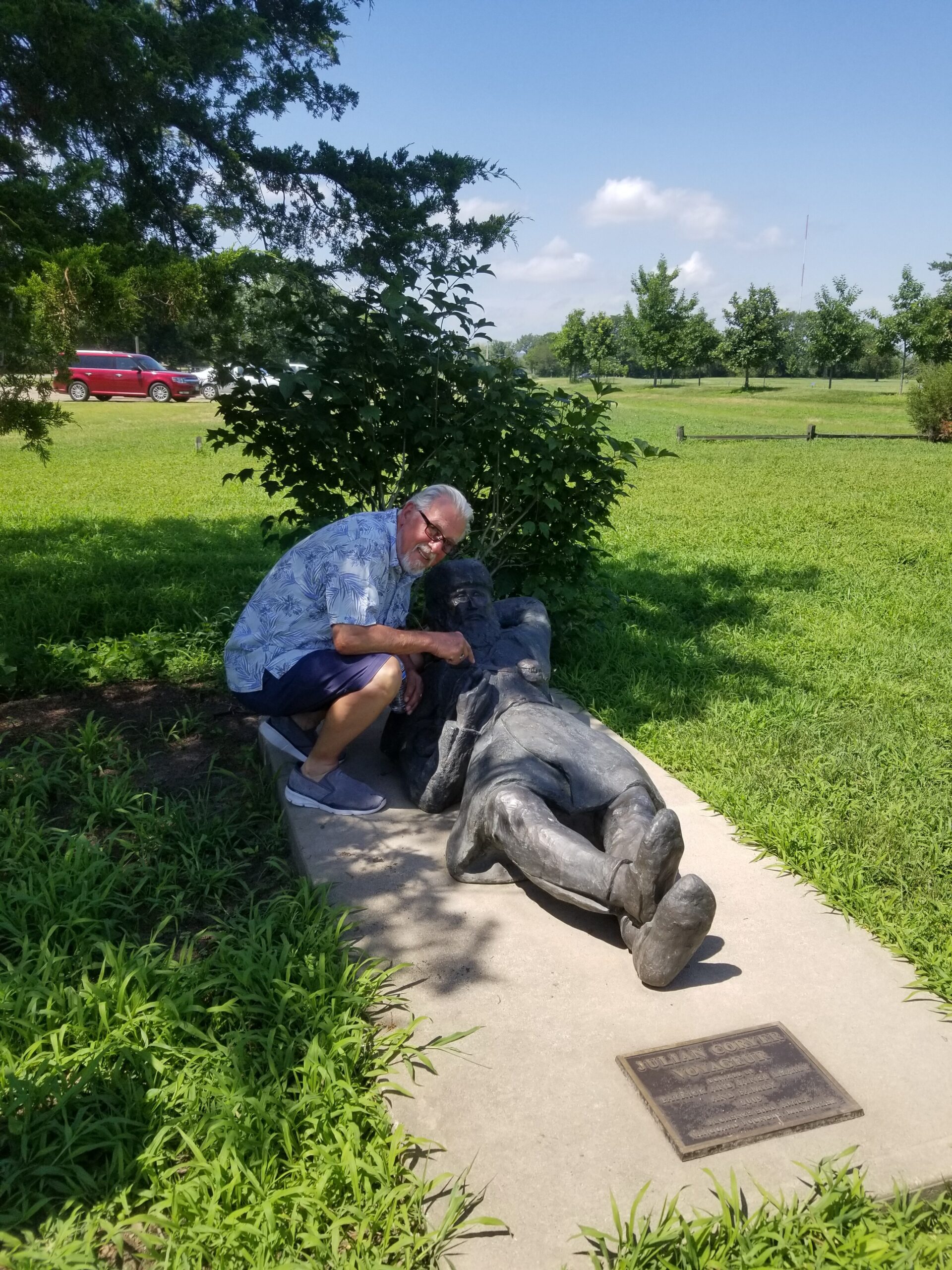 Older man in a blue floral shirt kneels beside a reclining bronze statue along a park path, with a plaque nearby.