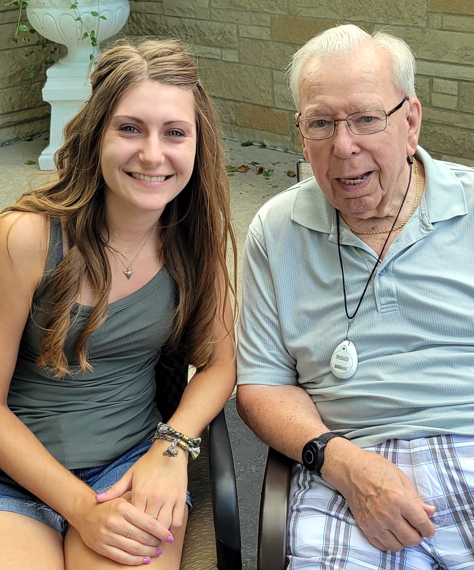 Smiling young woman in a green tank top sits beside an elderly man with glasses on a bench outdoors.