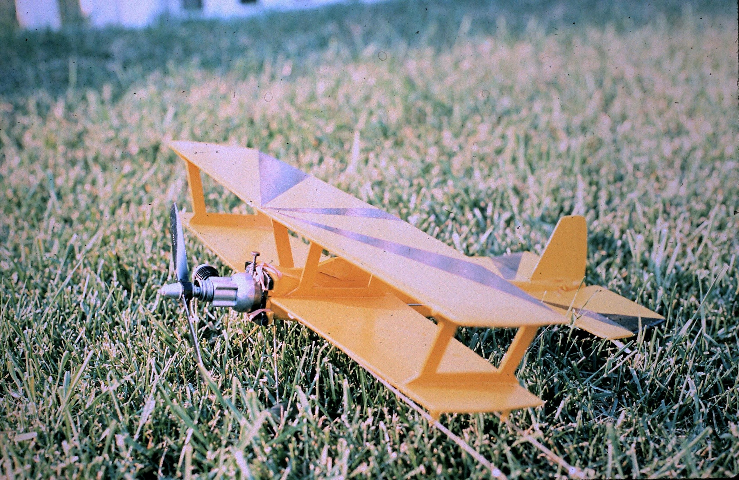 Small yellow model airplane resting on a grassy field, visible engine at the front and propeller bar extending forward.
