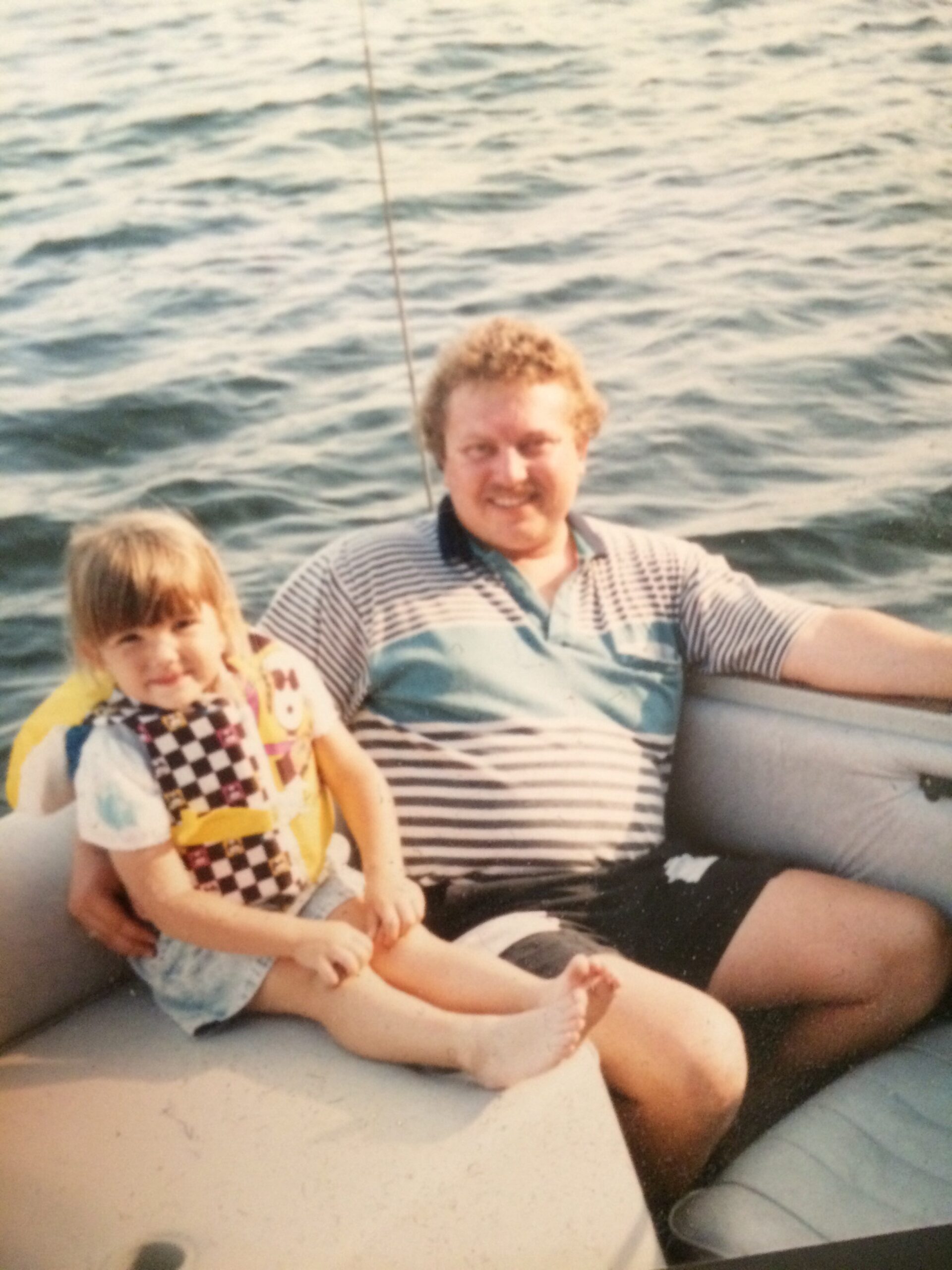Man with striped polo sitting on a boat next to a smiling young girl in a colorful vest, open water behind them.