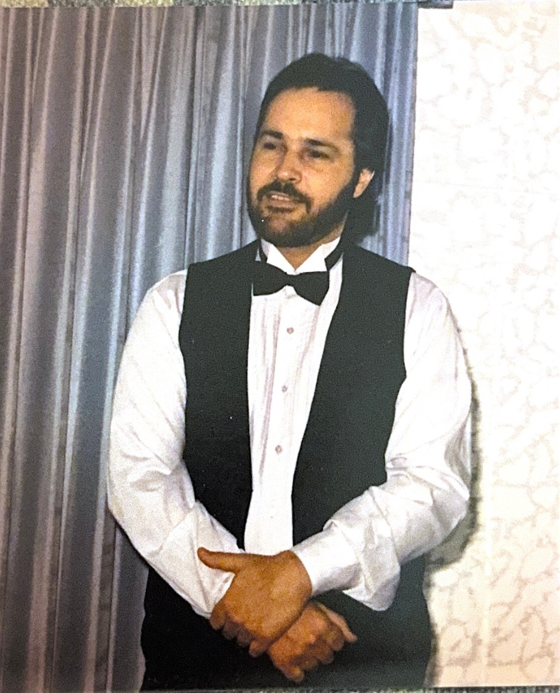Man with dark hair and beard wearing a white shirt, black vest, and bow tie, standing with arms crossed in front of a curtain.