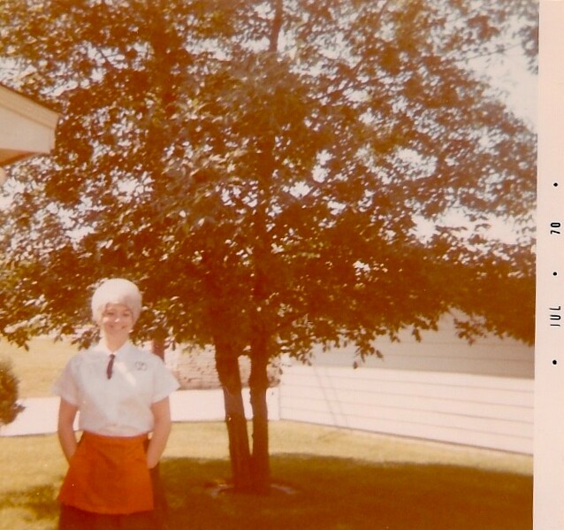 Woman with short white hair in a white blouse and red skirt standing on a lawn beside a tree in a sunny yard.