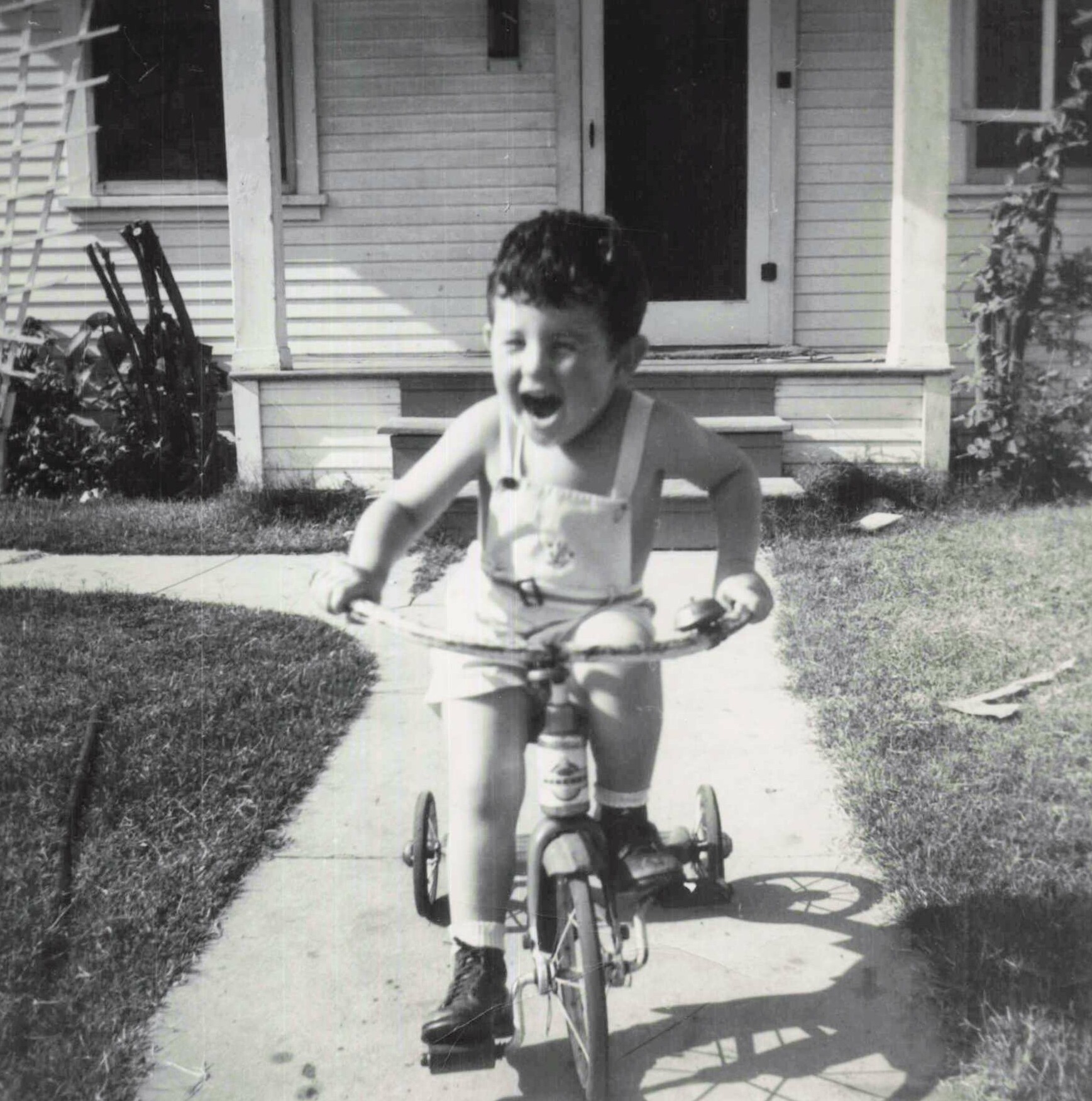 Boy rides a tricycle on the sidewalk in front of a house, mouth open as if laughing or shouting.