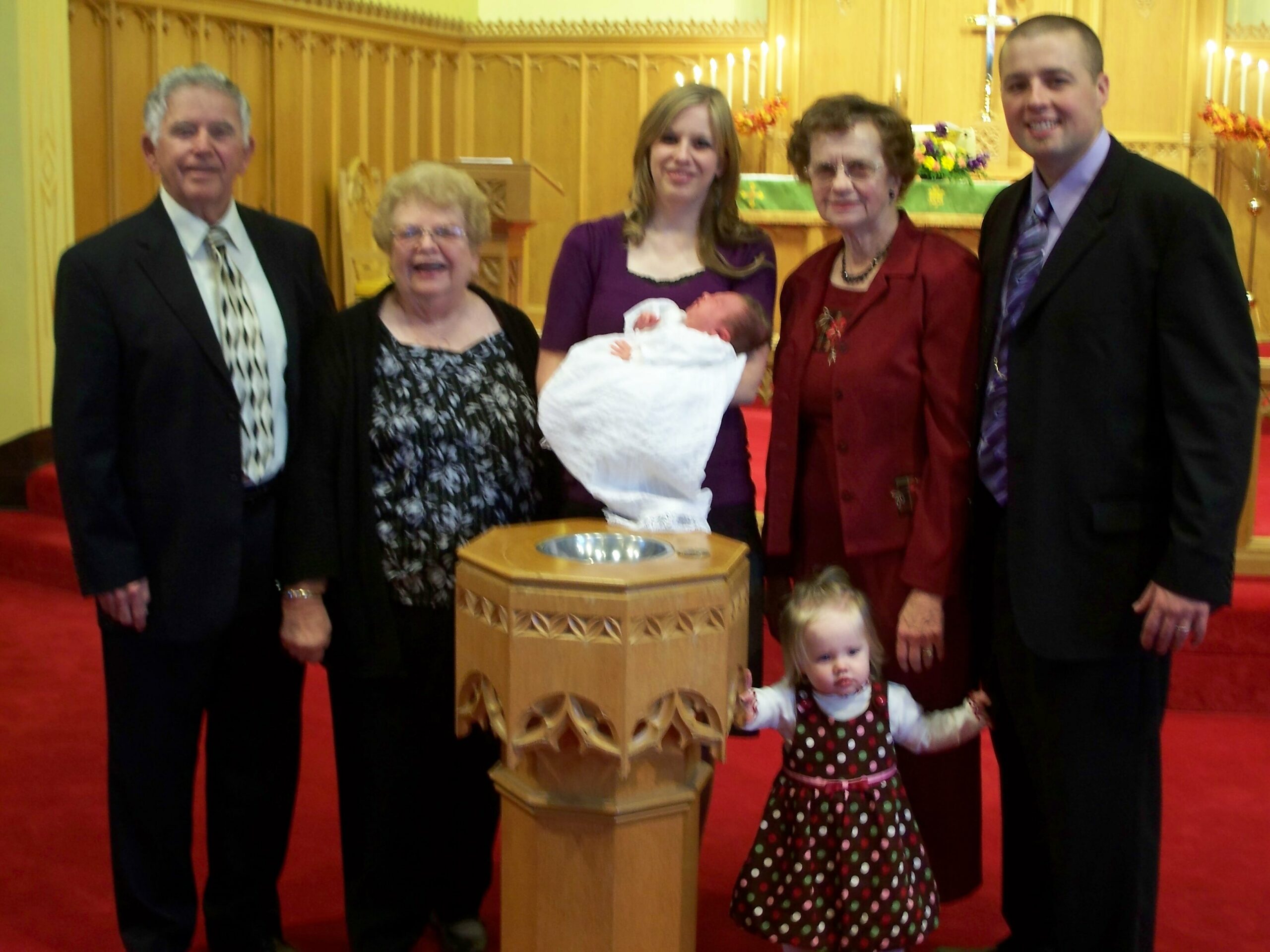 Six people posing in a church, with a newborn baby wrapped in white held by a woman in purple at the center, and a small child in a polka-dot dress in front; warm wood altar and candles in the background.