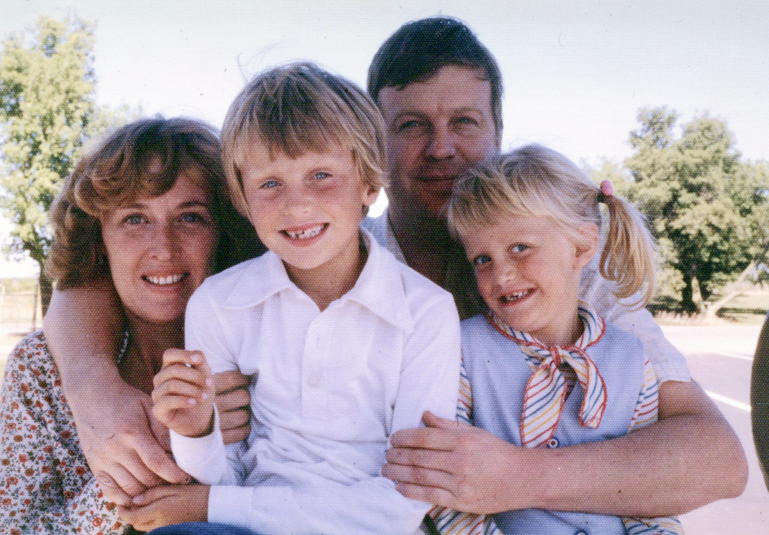 A family of four posing closely together outdoors on a sunny day, smiling at the camera (mother on left, father behind them, boy center, girl on the right).