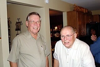 Two older men smiling at a home gathering, standing in a kitchen area.