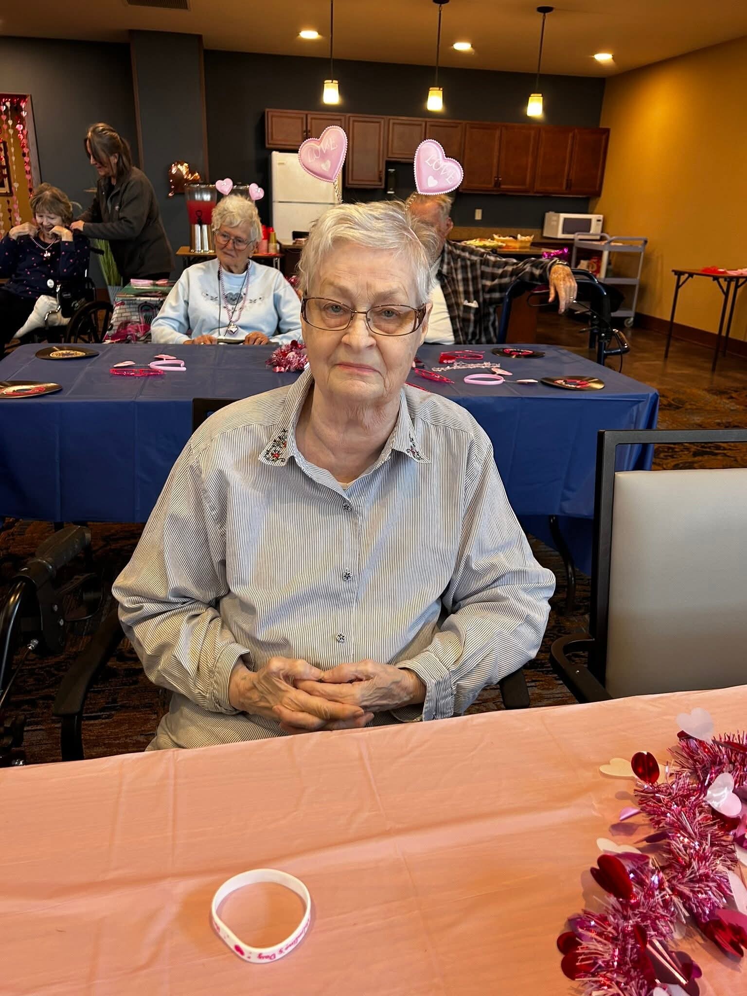 Elderly woman with glasses sits at a pink‑themed party table, with heart decorations in the background.