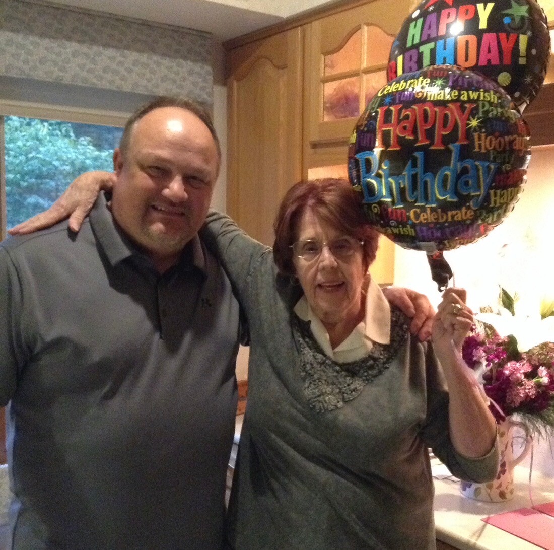 Man and elderly woman pose together in a kitchen with a colorful 'Happy Birthday' balloon beside them in celebration.