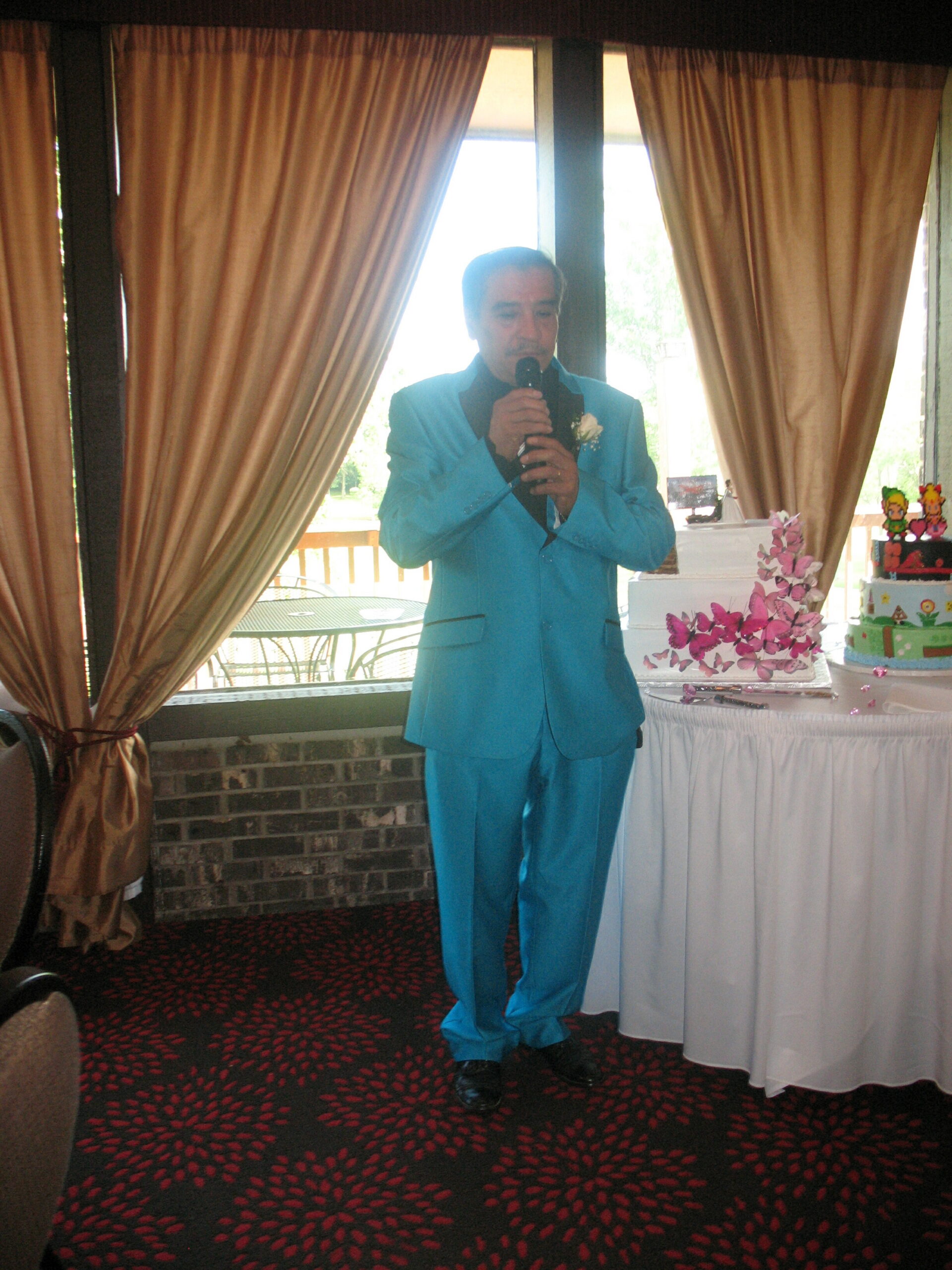 Man in a bright turquoise suit holding a microphone at a party near a decorated cake display.