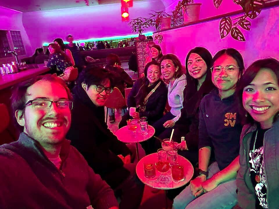 Group of friends seated around two small tables at a bar, smiling under pink neon lighting with plants on the wall.