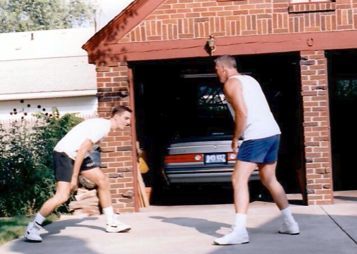 Two men playing basketball on a driveway in front of a brick garage; a car is visible inside the open garage.