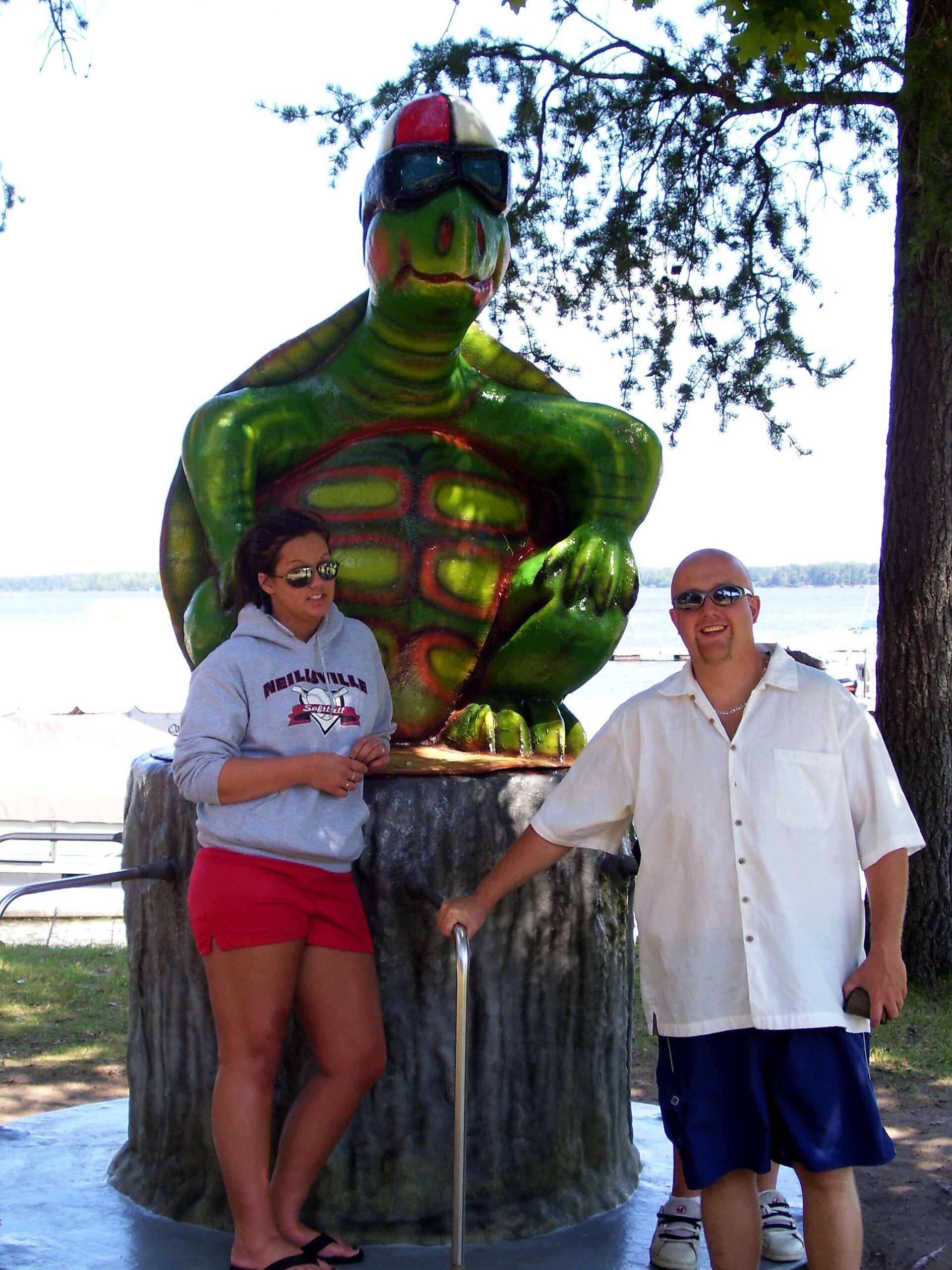 Two people posing beside a large green dinosaur statue wearing sunglasses outdoors by a lake.