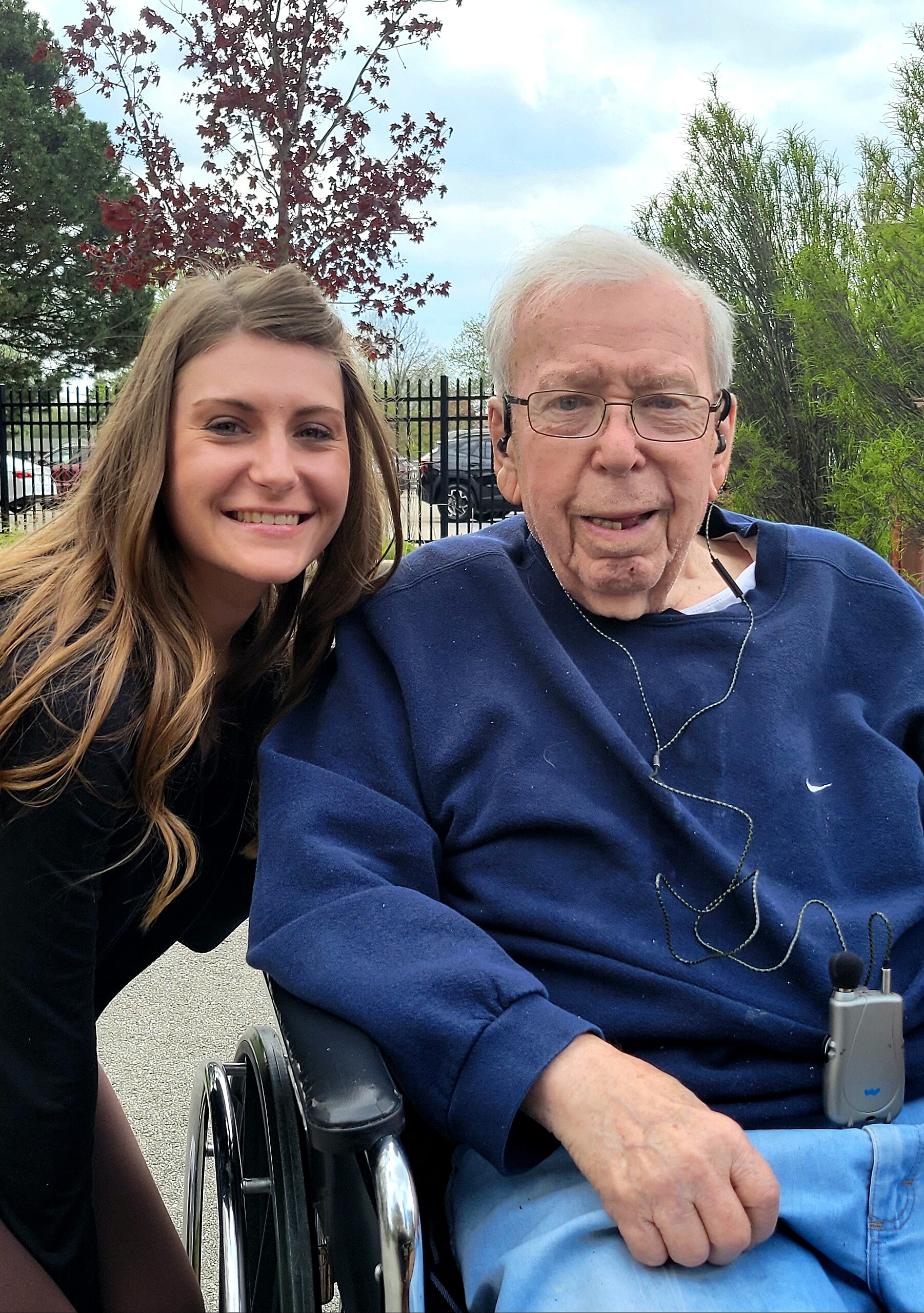 Smiling young woman beside an elderly man in a wheelchair outdoors, with a fence and trees in the background.