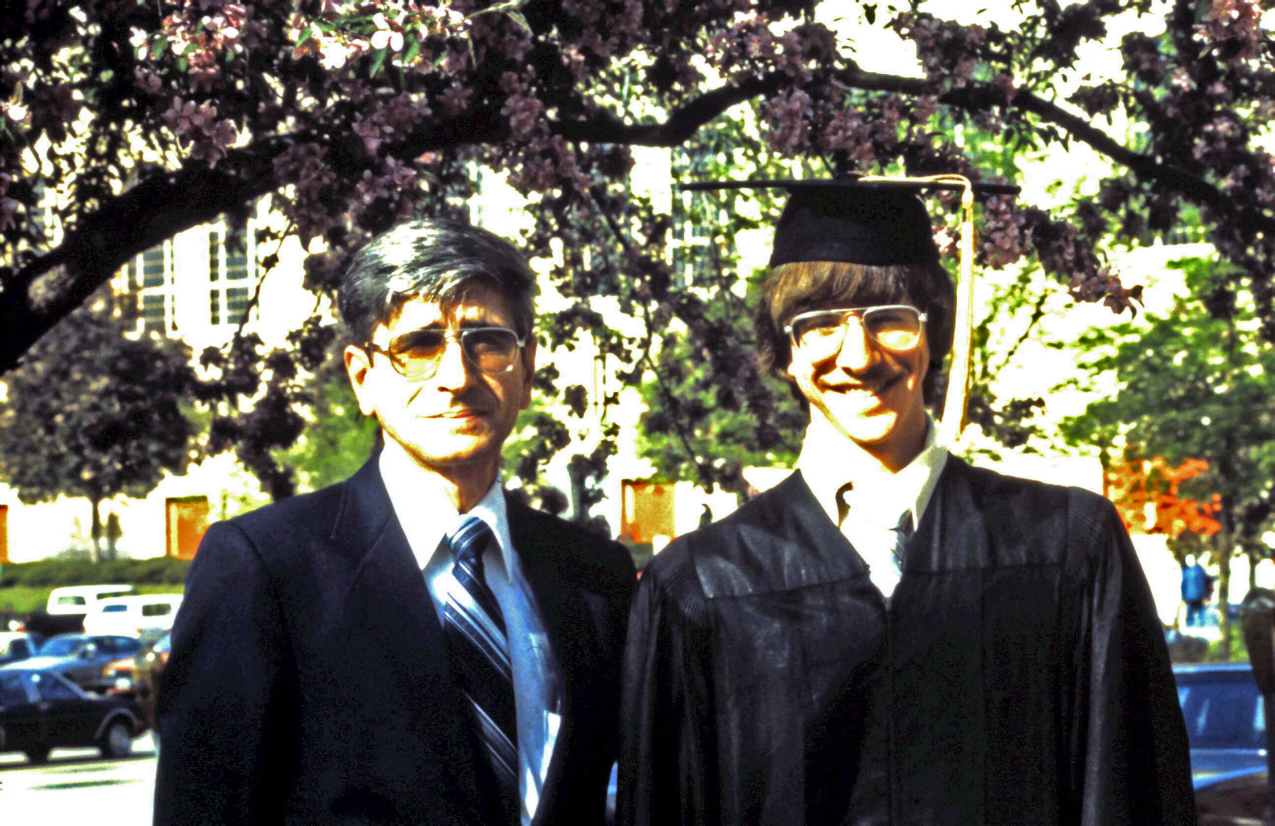 Two men in suits posing outdoors; one wears a graduation cap and gown while the other is in a suit.